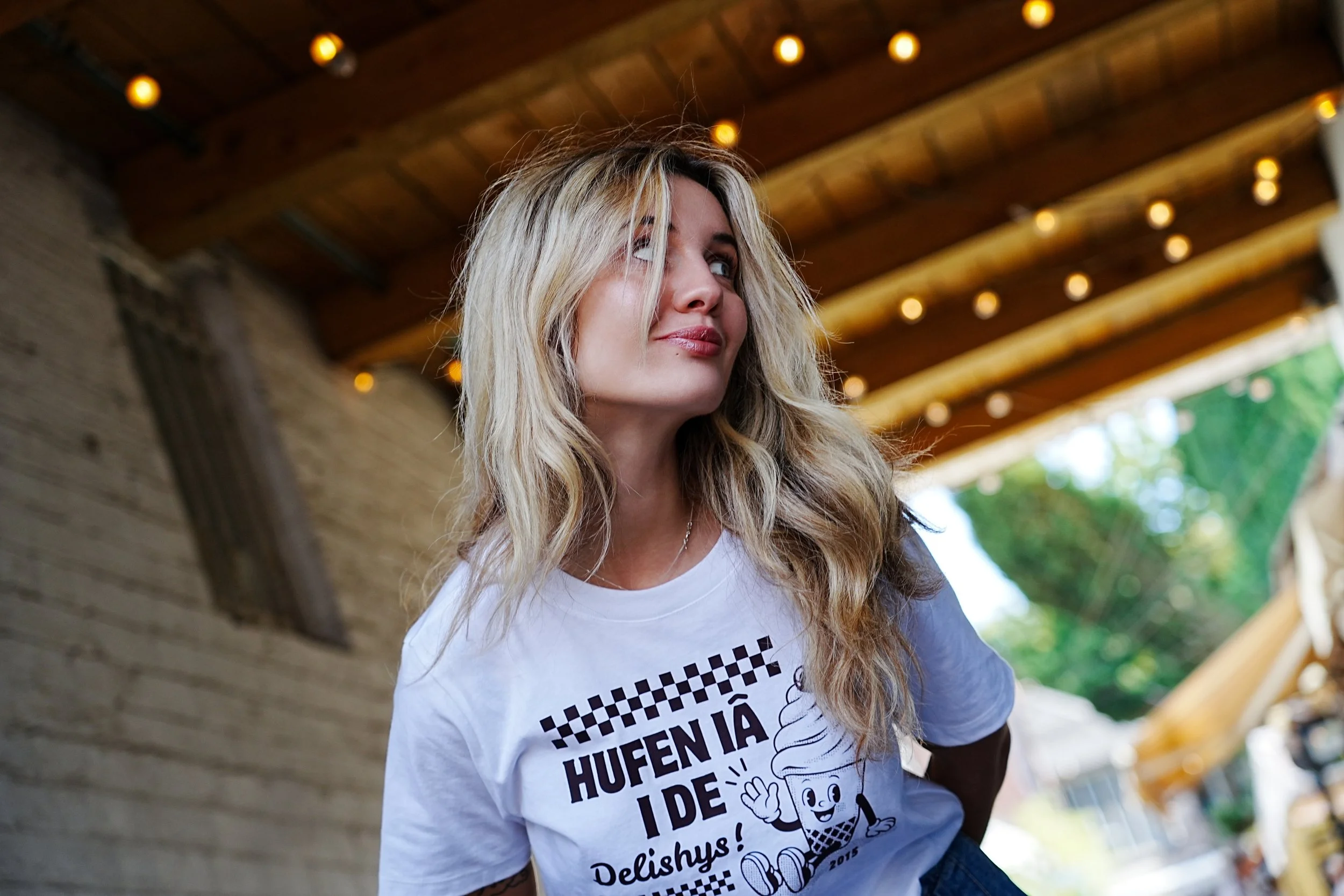 A blonde woman with wavy hair wearing a white T-shirt with Hufen Ia on it.. She is outdoors under a wooden ceiling with string lights, smiling and looking to the side. This is a very cool way to show a welsh language clothing brand
