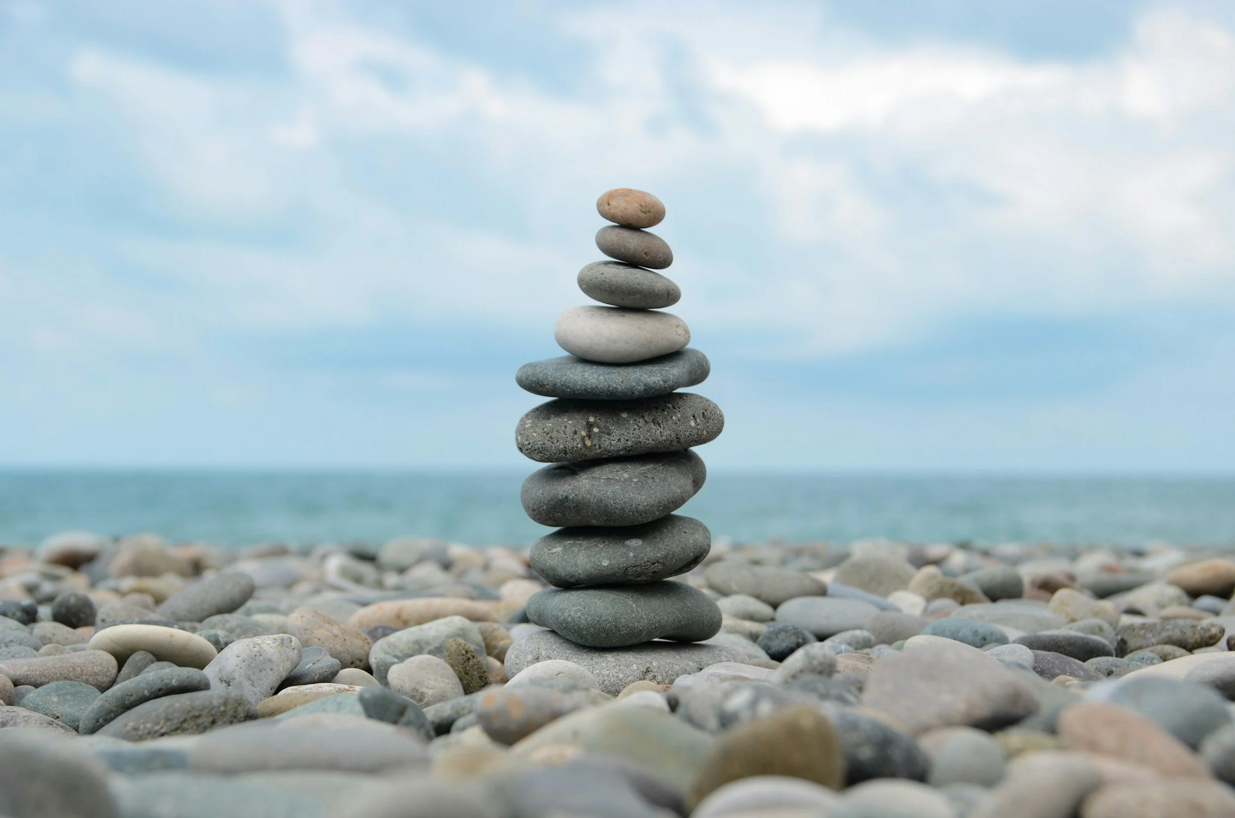 Stacked smooth stones of various sizes and colors on a pebble-covered beach with a cloudy sky and ocean in the background.