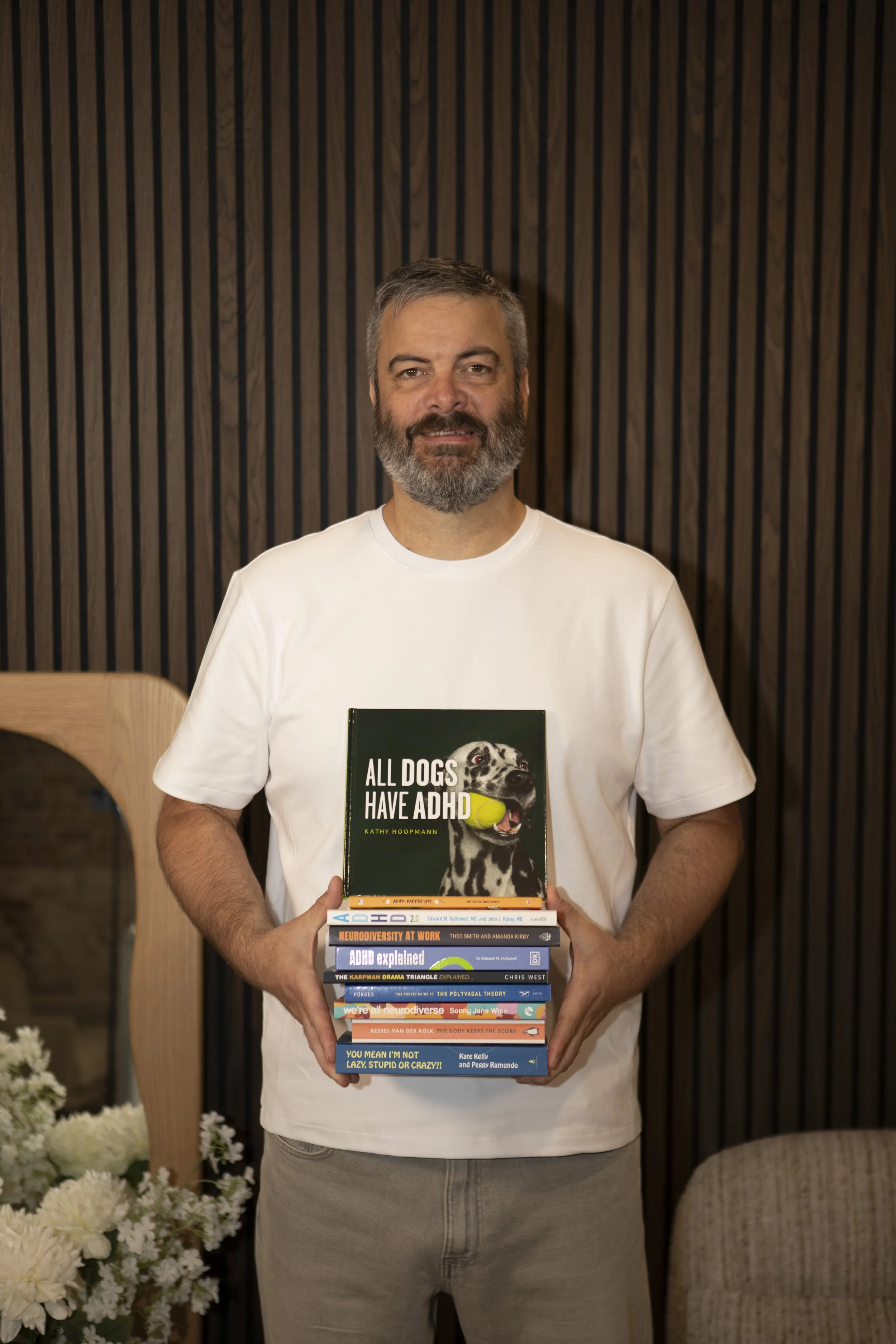 Bearded man in a white T-shirt holds a stack of books about ADHD, neurodiversity, and mental health.