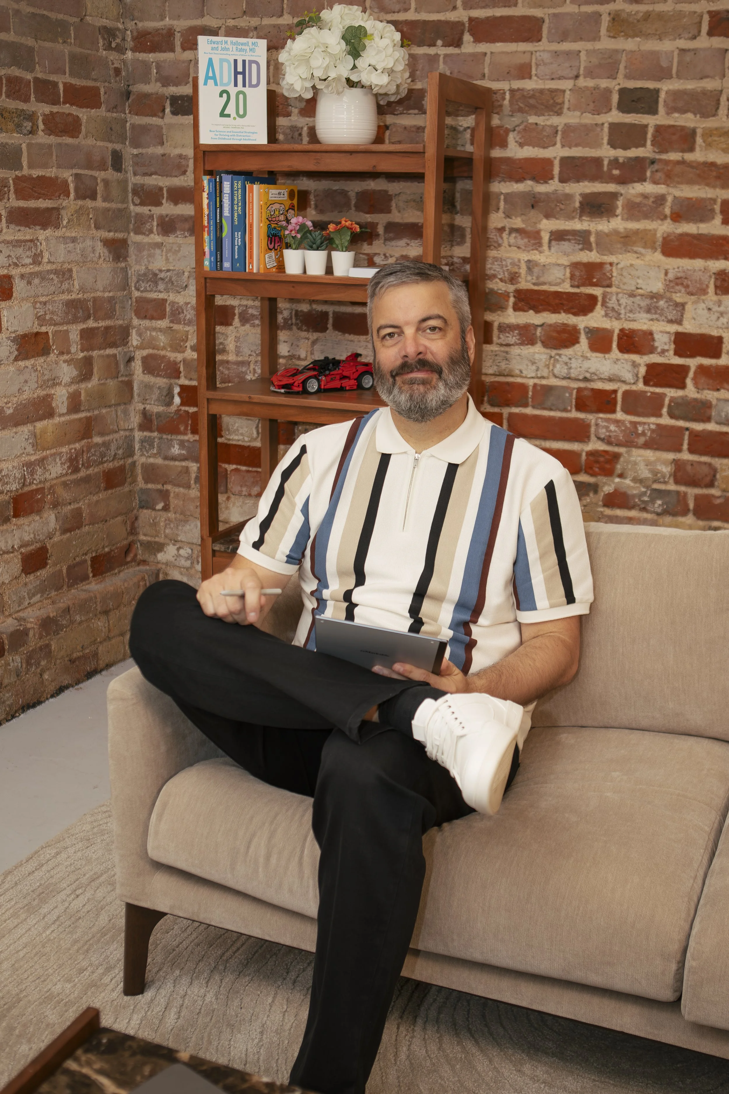 A man with gray hair and a beard sitting on a beige couch in front of a brick wall. He is holding a tablet, wearing a striped polo shirt and black pants with white sneakers. Behind him is a wooden shelf with books, potted plants, a white vase with hydrangeas, and a red toy car.