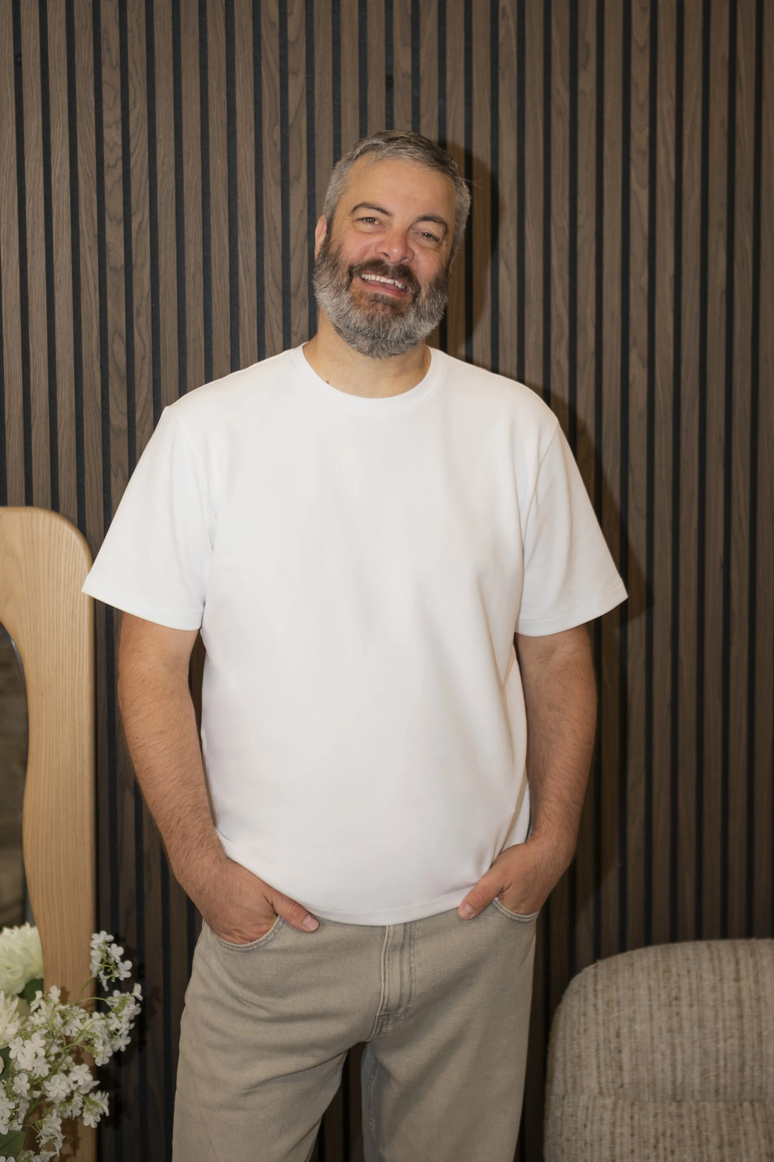 A middle-aged man with a beard and gray hair, smiling, wearing a plain white t-shirt and beige pants, standing in front of a wood-paneled wall with a small chair and white flowers nearby.