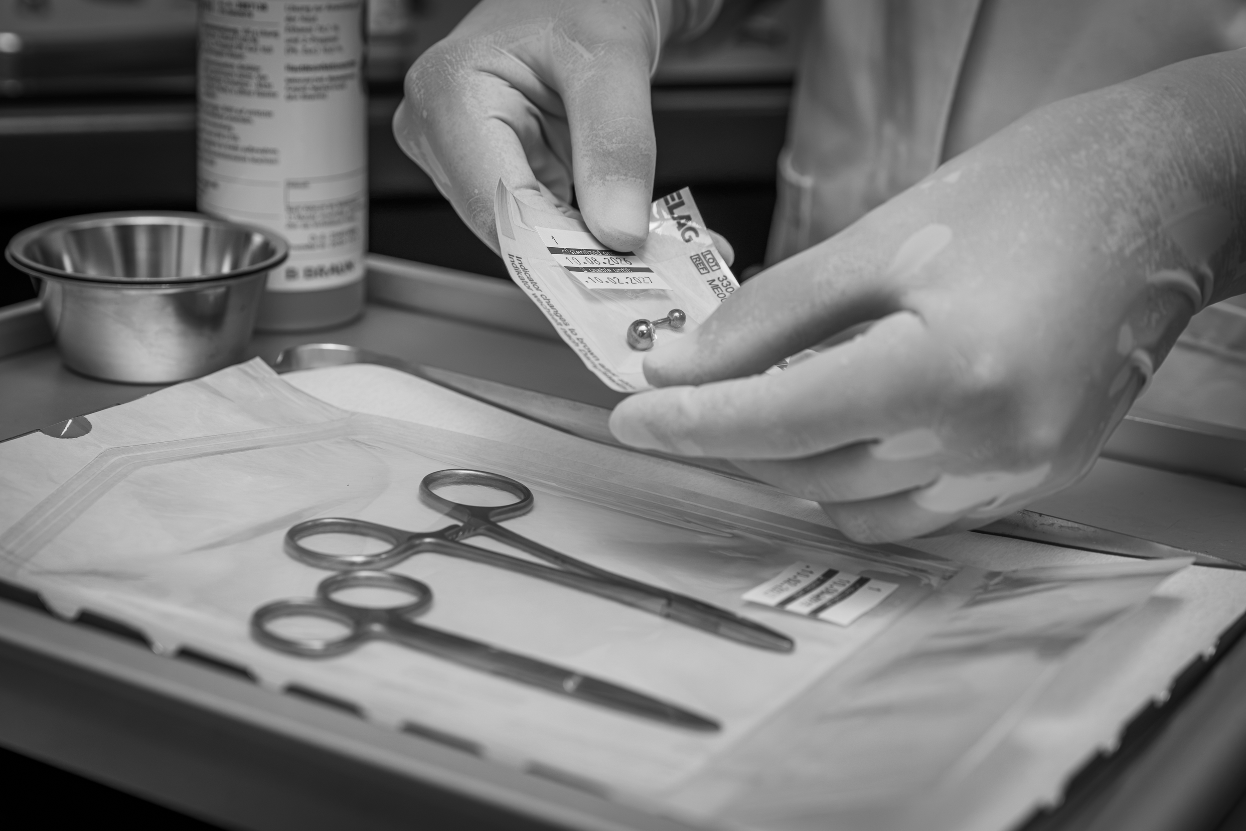 Eine Hand in einem medizinischen Handschuh hält eine Verpackung mit einer medizinischen Spritze. Auf dem Tisch liegen medizinische Instrumente wie Scheren und chirurgische Klammern. Im Hintergrund sind medizinische Behälter und Flaschen sichtbar.