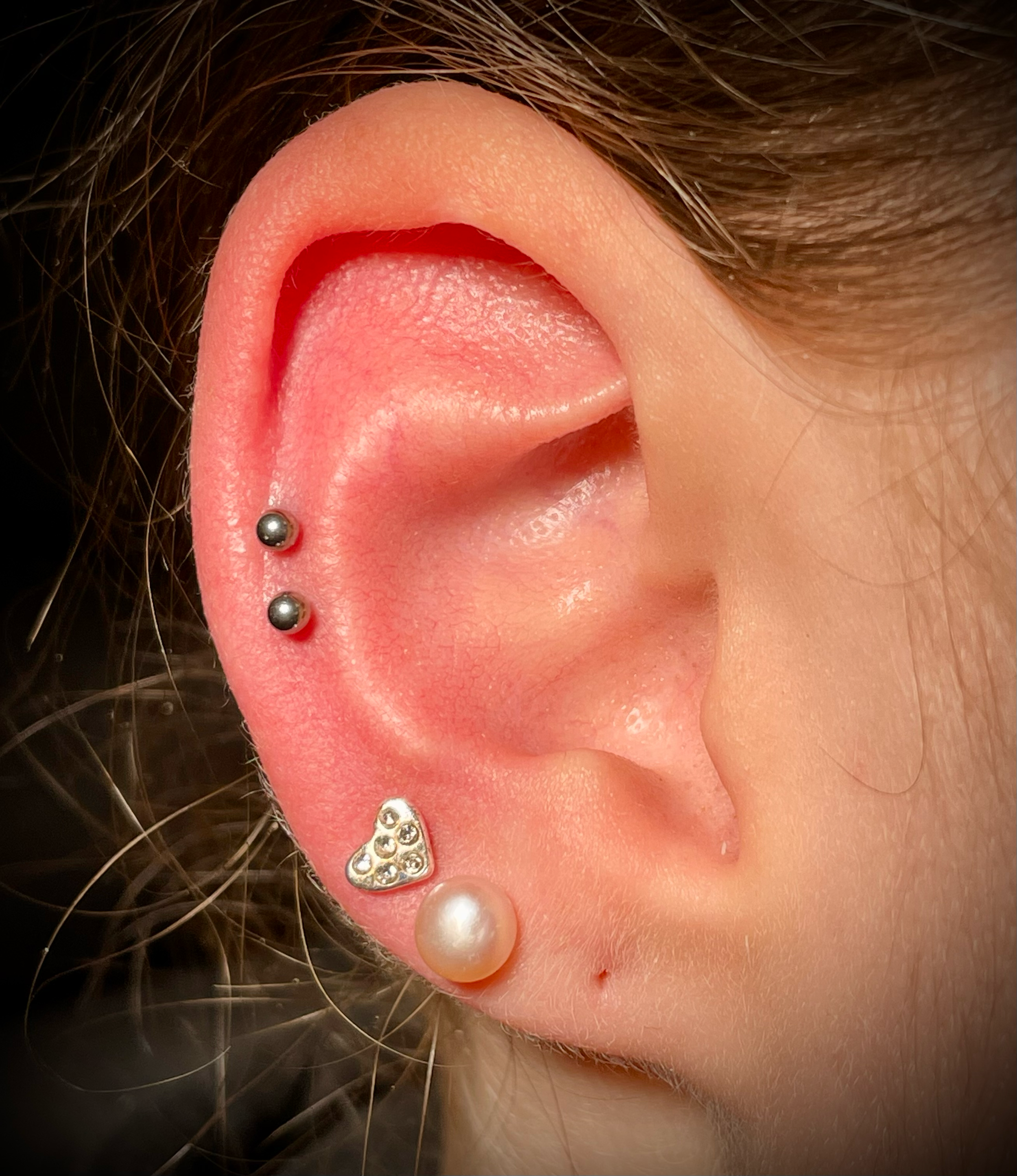 Naheaufnahme eines weiblichen Ohrs mit mehreren Piercings und Schmuck, umgeben von dunklem Haar.