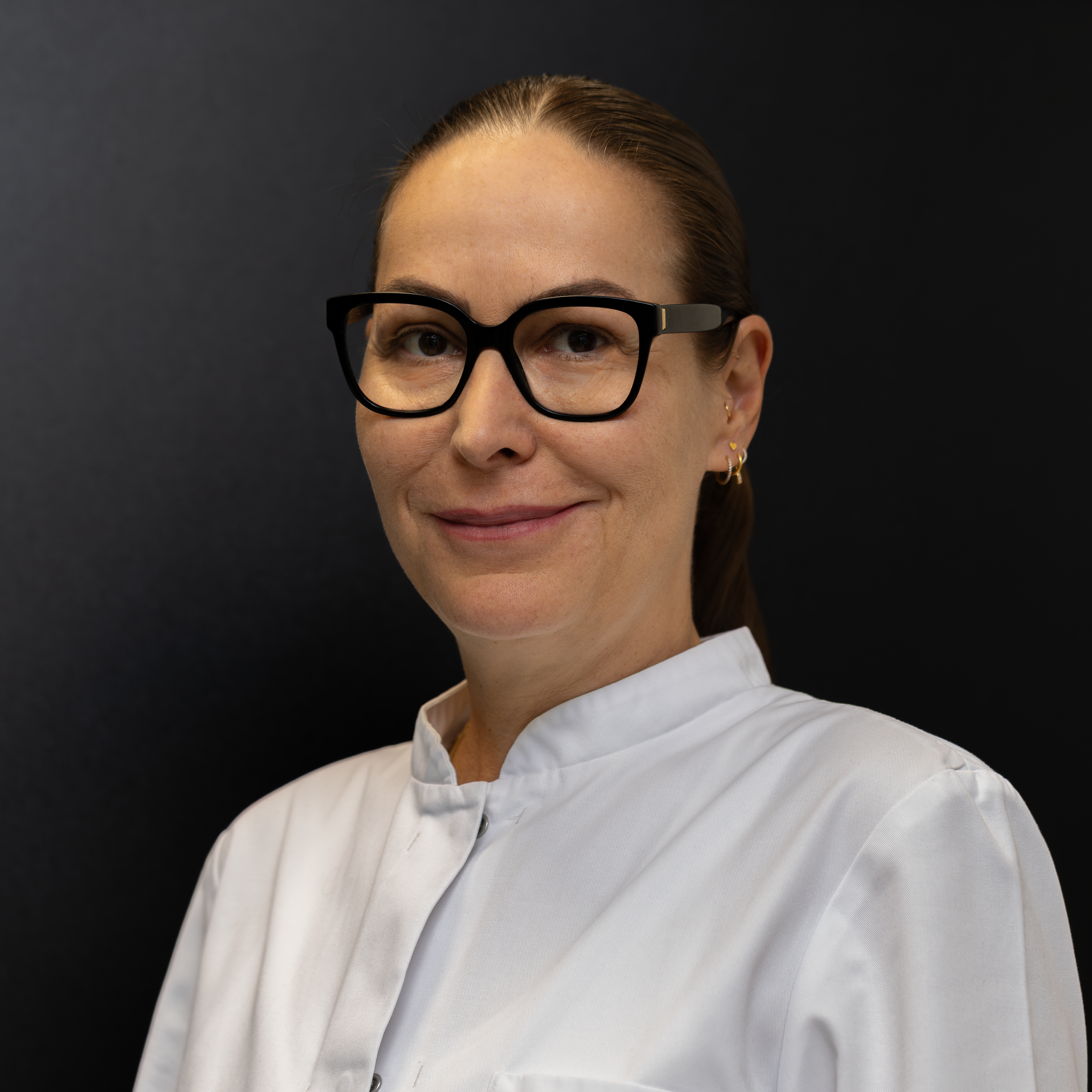 Porträt einer Frau mit braunen Haaren, schwarzen Brillen und mehreren Ohrsteckern, trägt einen weißen Arztkittel vor einem schwarzen Hintergrund.