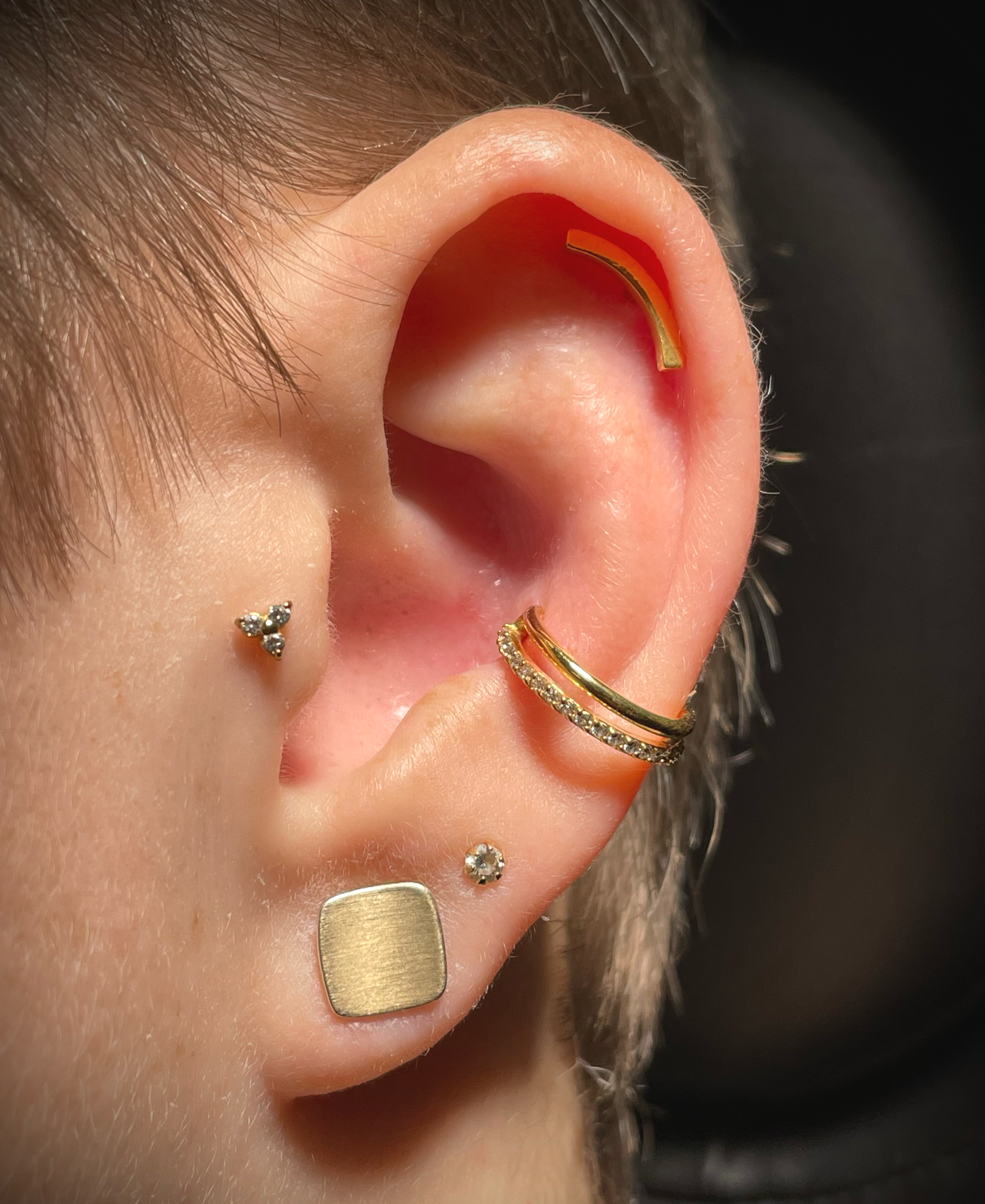 Nahaufnahme eines Ohres mit mehreren Ohrringen, darunter ein goldener Gehörknorpelschmuck, ein doppelter goldener Ring mit Kristallen und ein quadratischer goldener Ohrring. Haare und Haut sind sichtbar.