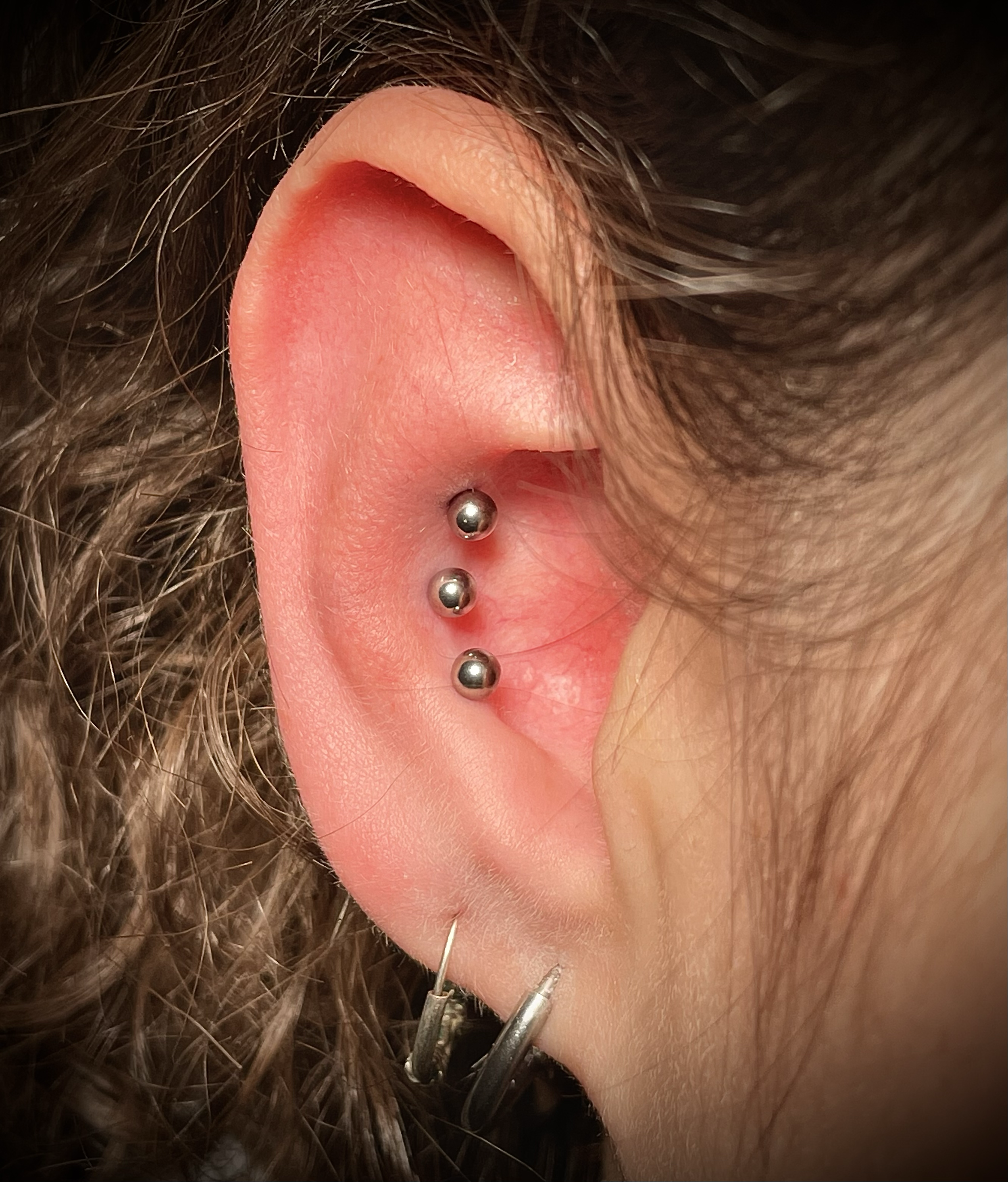 Nahaufnahme eines menschlichen Ohres mit mehreren Piercings, darunter ein Piercing mit mehreren Kugeln an der Ohrläppchen und Helix, umgeben von dunklem, gewelltem Haar.