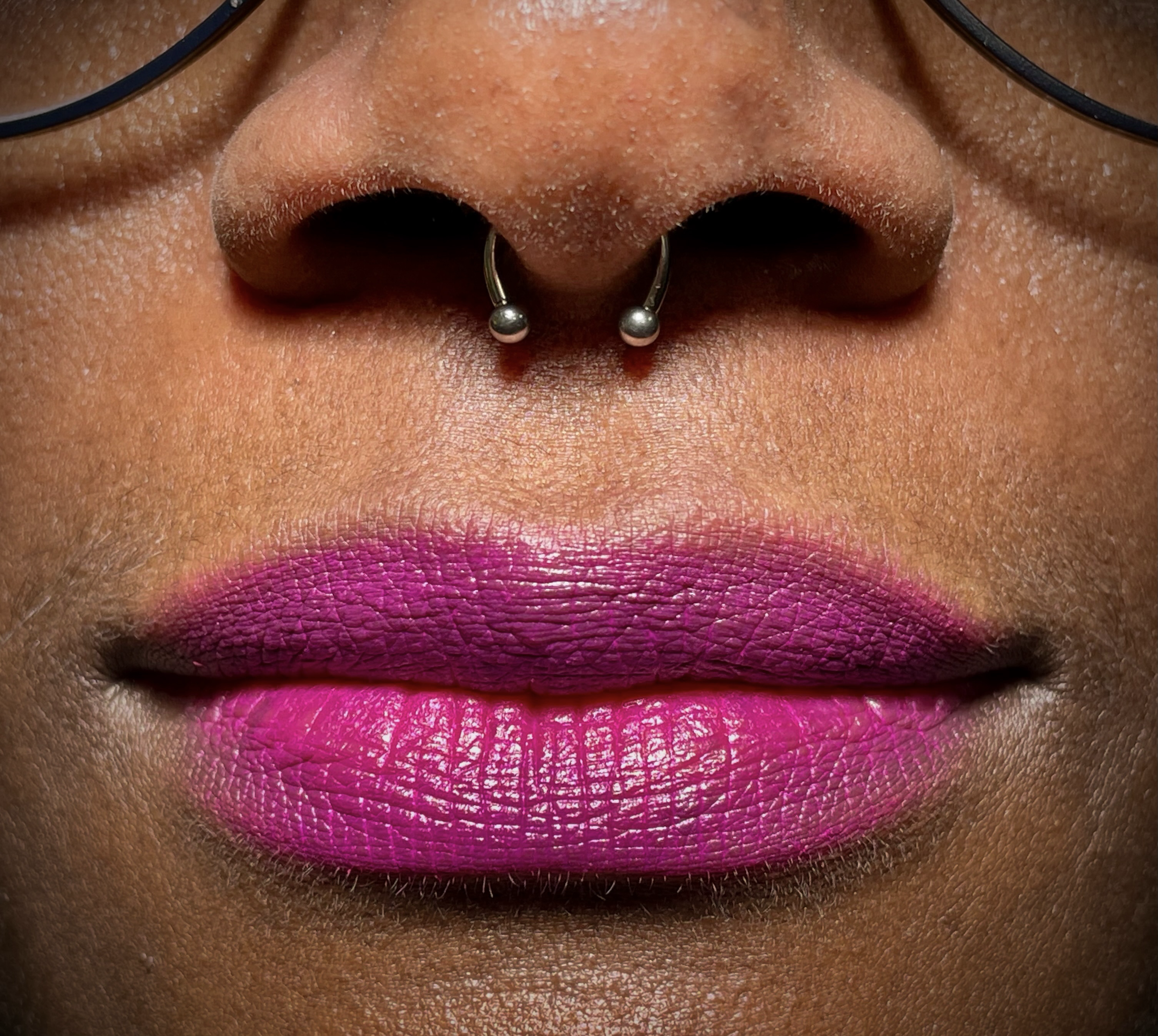 Nahaufnahme von Lippen mit pinkem Lippenstift und einer Nase mit Nasenring, Person trägt eine schwarze Brille.