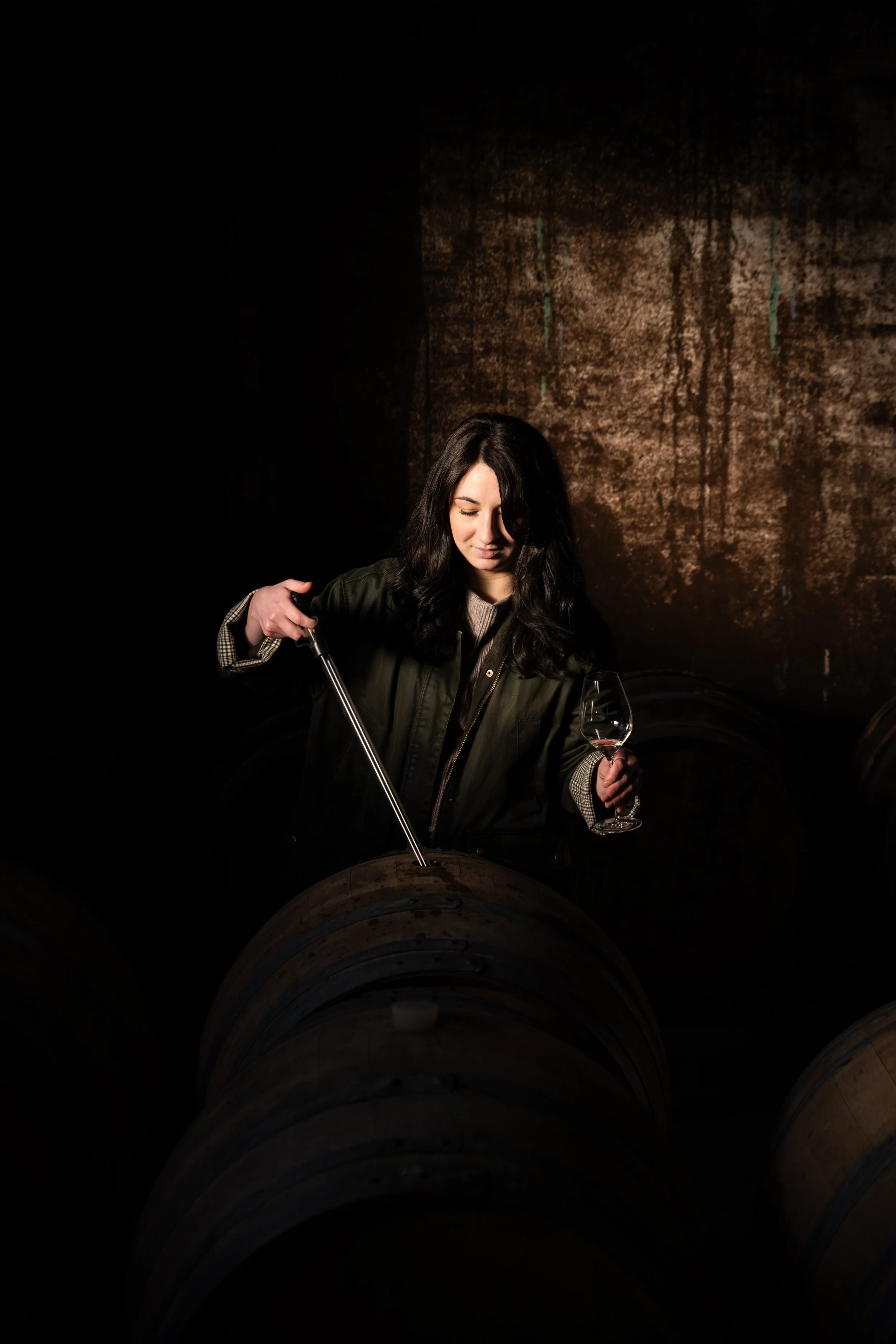 Une femme avec des cheveux longs noirs, portant une veste verte foncée, tient un verre de vin rosé dans une main et un tire-bouchon dans l'autre, dans un environnement sombre avec un mur en pierre en arrière-plan.