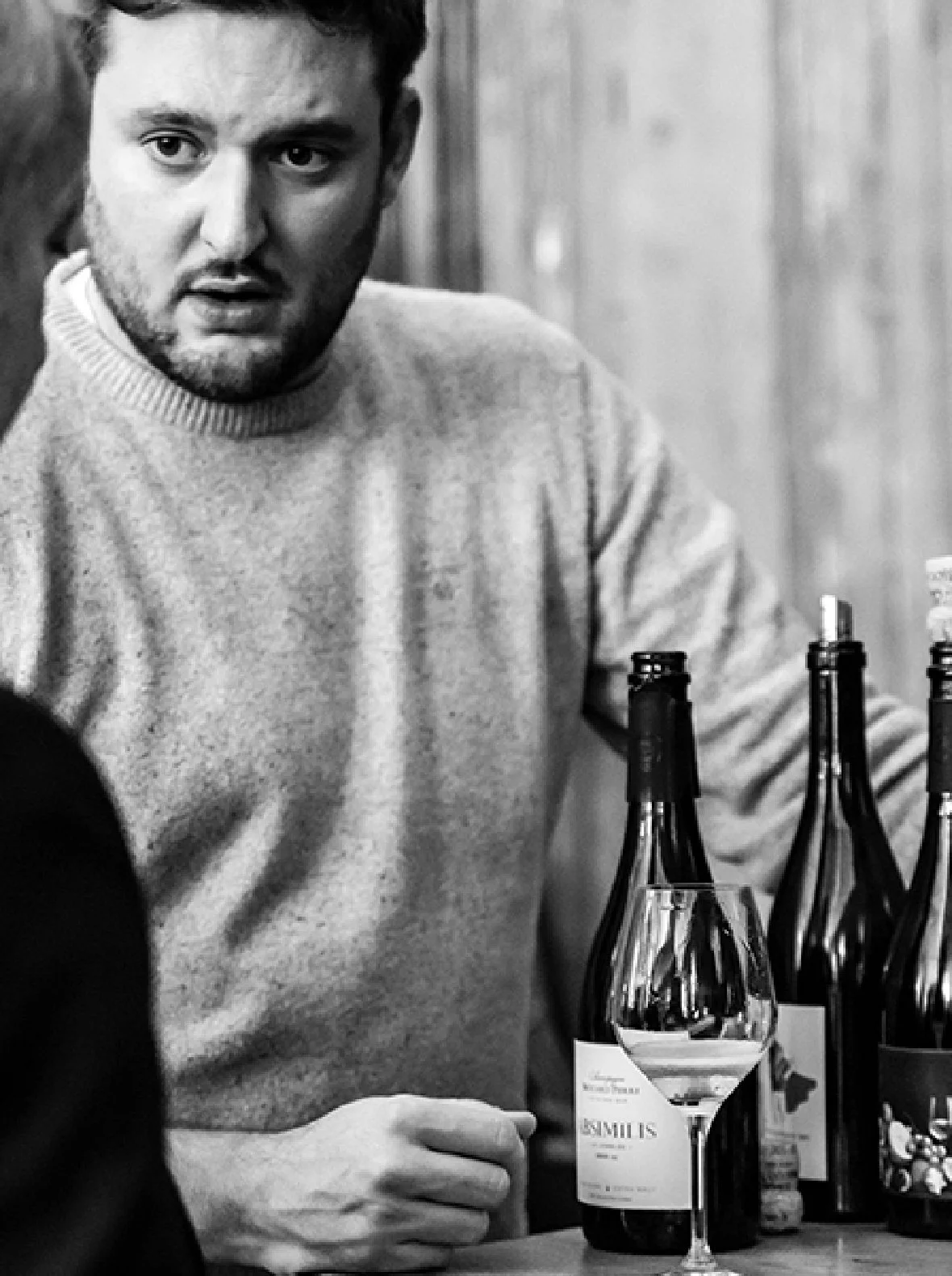 Un homme avec une barbe et un pull gris assis à une table, entouré de plusieurs bouteilles de vin et un verre de vin blanc. La photo est en noir et blanc.