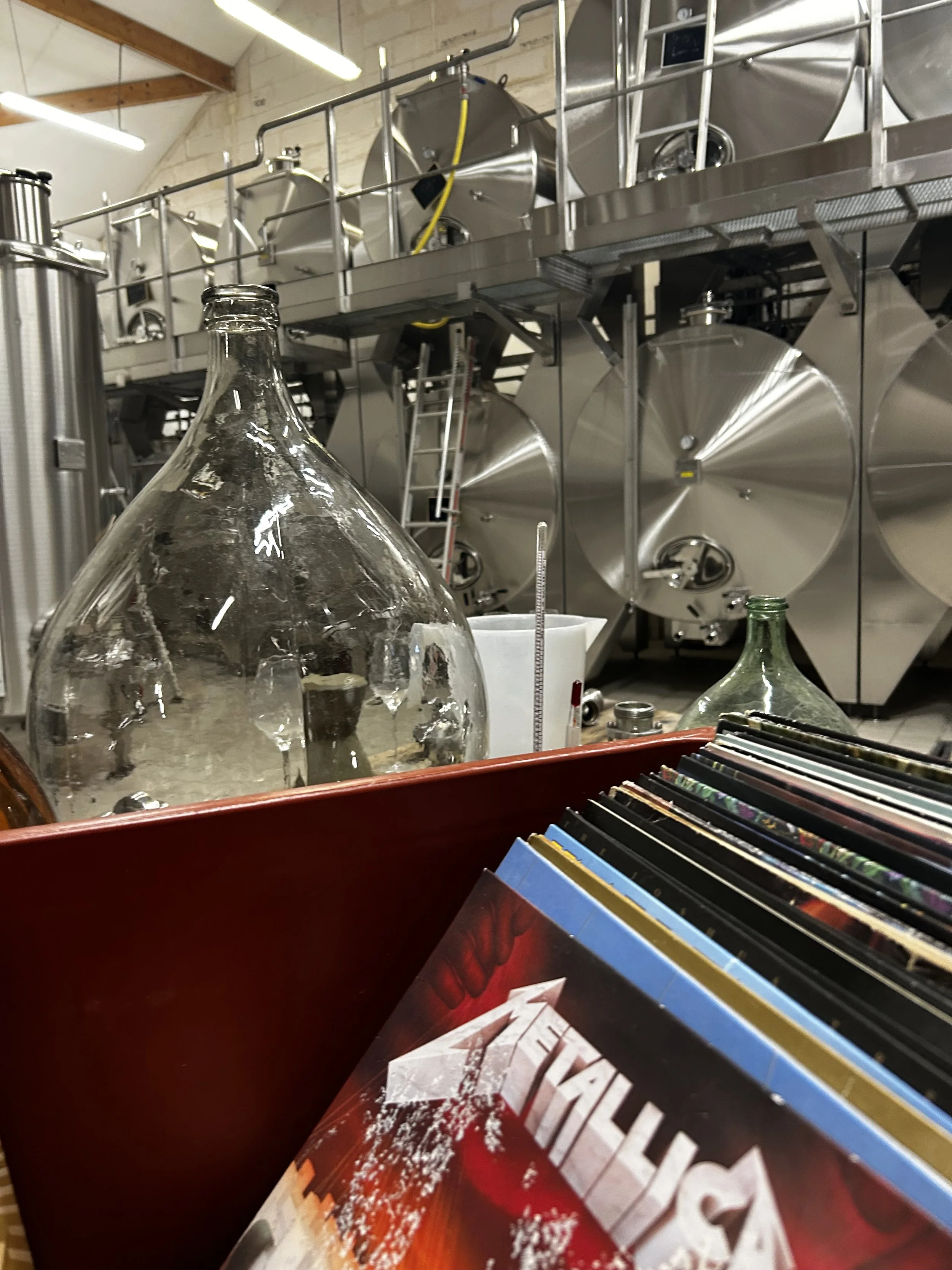 Photo d'une salle avec de grosses cuves en acier inoxydable utilisées pour la fabrication de bière ou de vin, avec une échelle en métal, et des boîtes de bandes dessinées ou livres dans le premier plan.