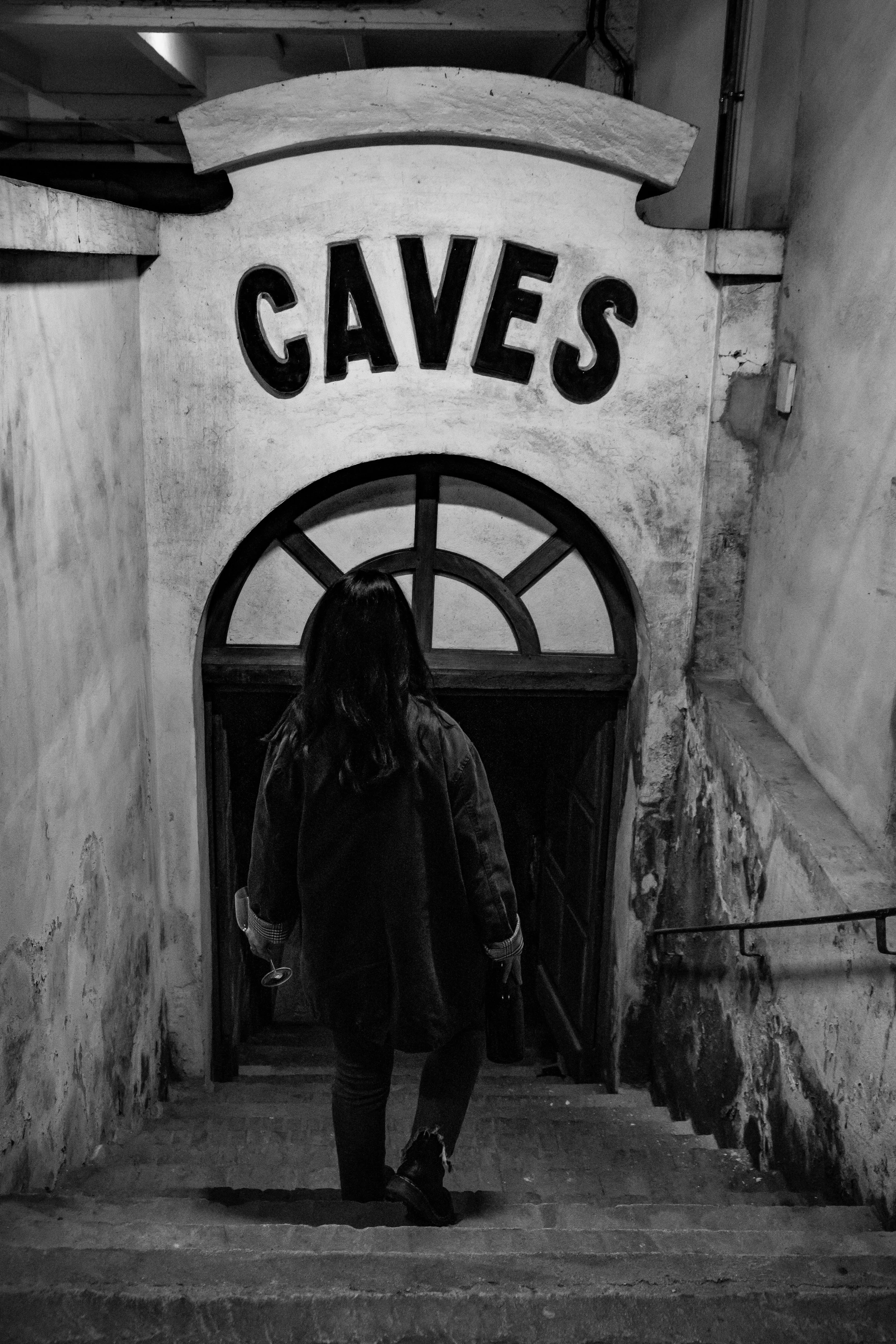 Une personne avec des cheveux longs portant un manteau noir descend un escalier dans une cave ancienne, avec un panneau indiquant 'CAVES'.