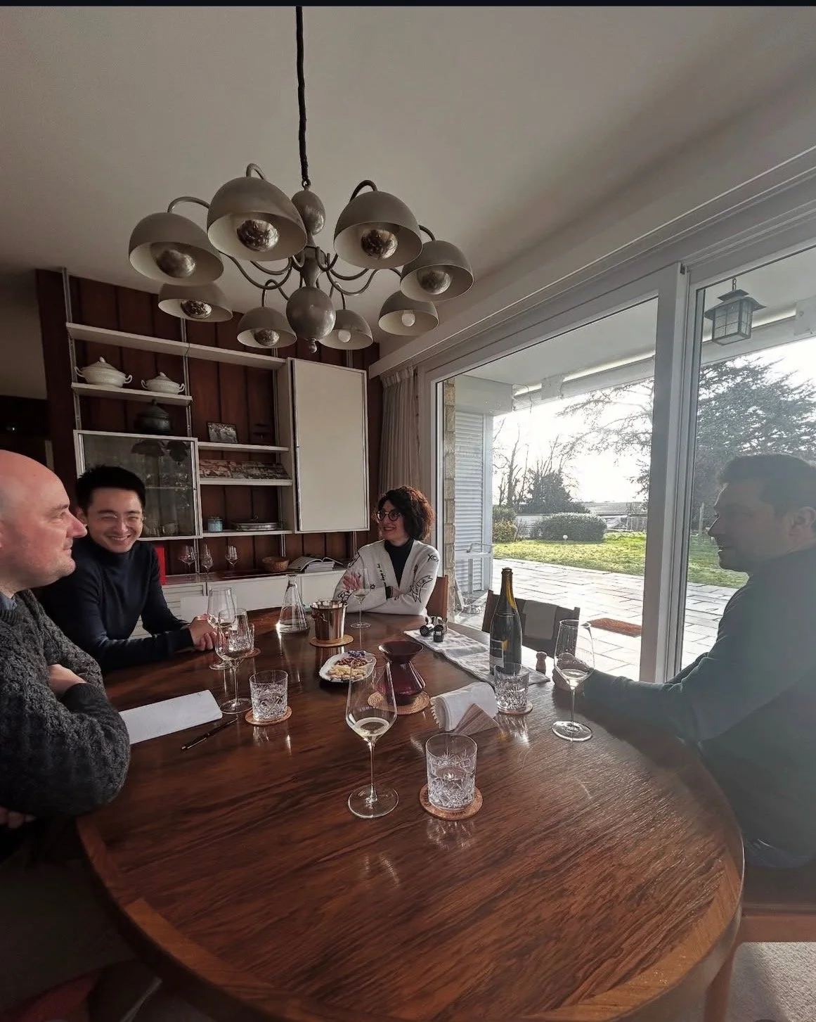 Quatre personnes assises autour d'une table en bois dans une salle bien éclairée, partageant un moment convivial avec des verres et une bouteille de vin, avec une vue extérieure par de grandes fenêtres.