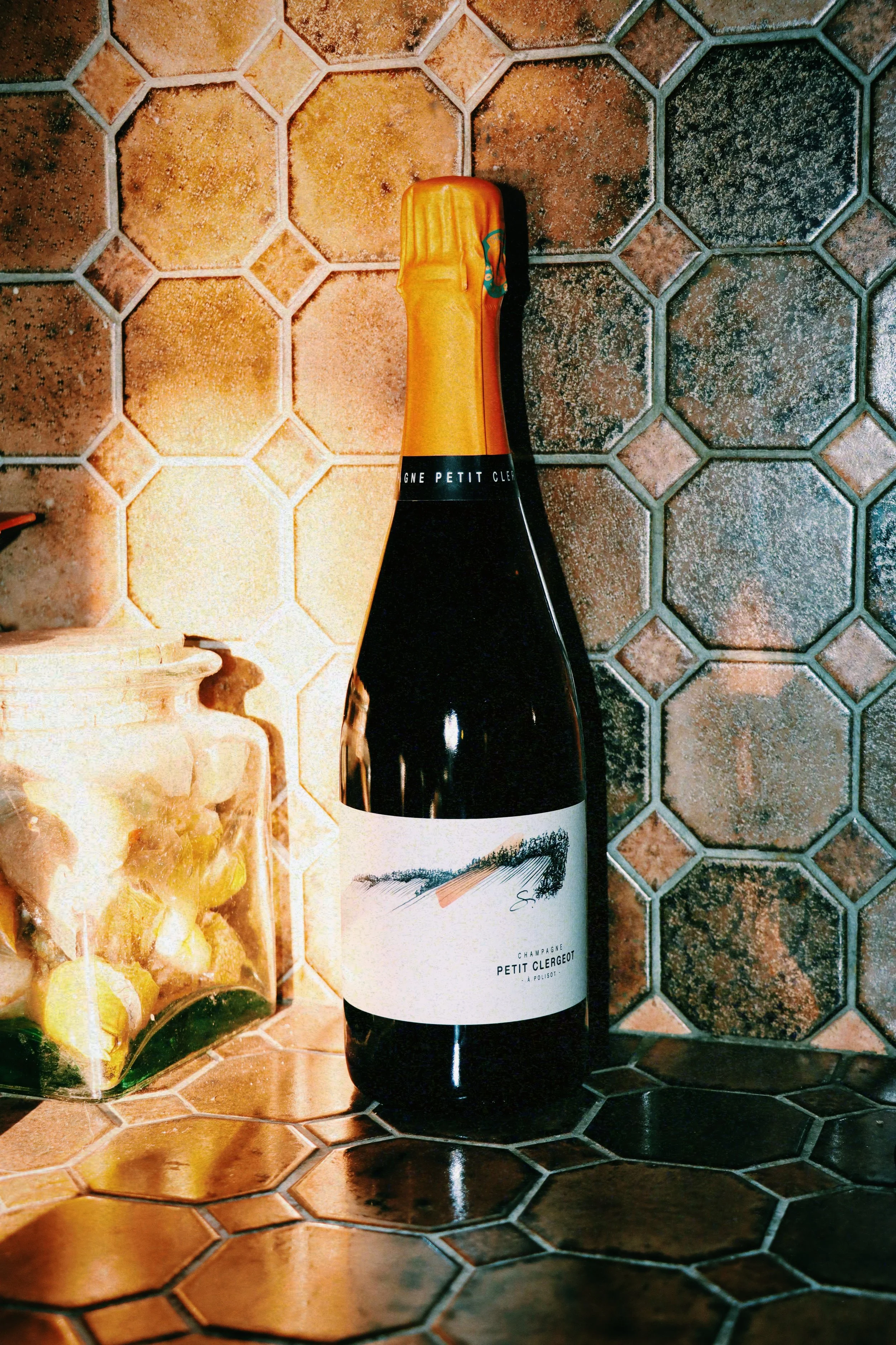 Une bouteille de champagne posé sur un plan de travail en carreaux de céramique hexagonaux bruns, à côté d'un pot en terre cuite.