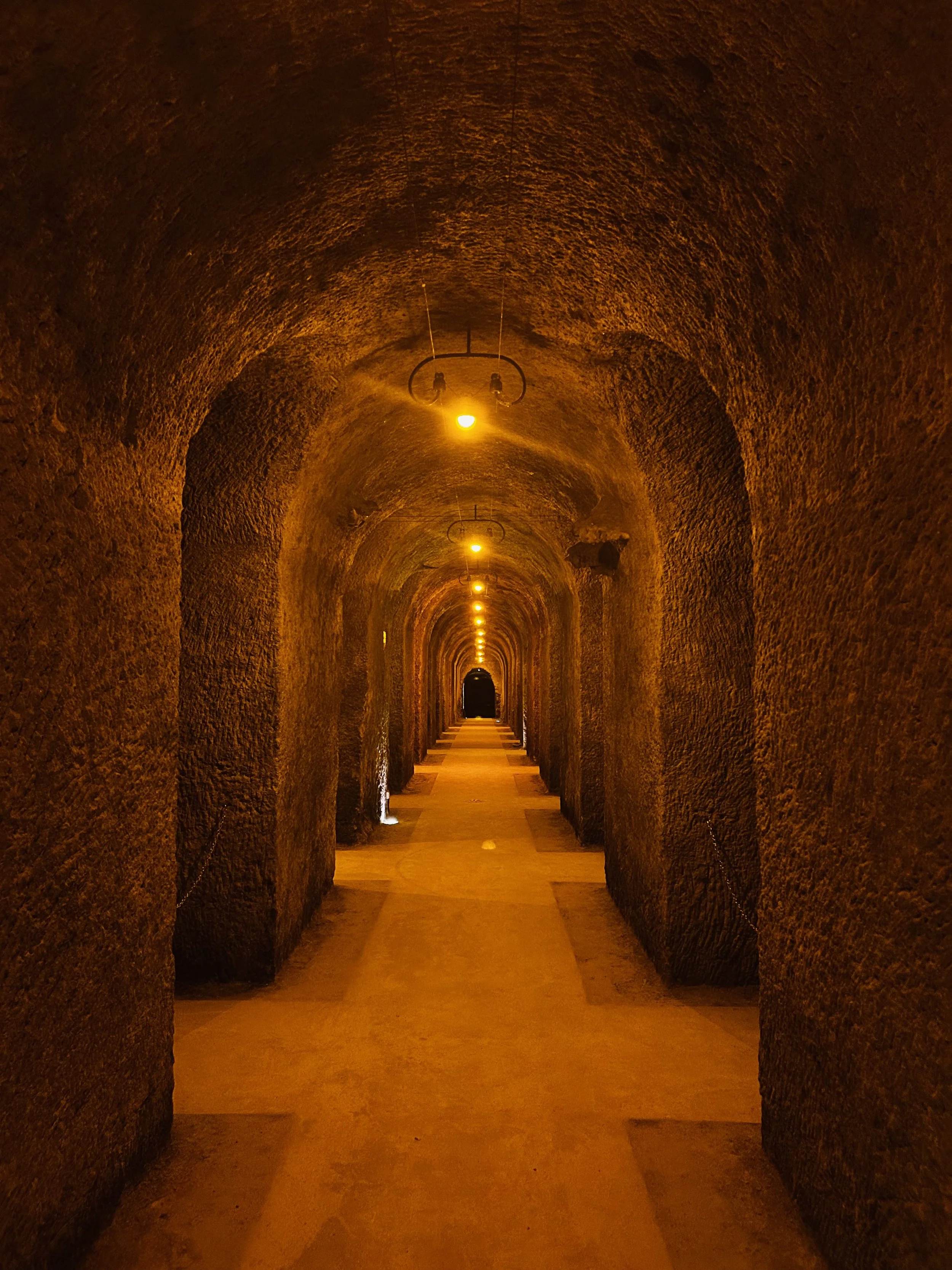 Longue galerie souterraine en pierre avec des arches et un éclairage jaune, vue jusqu'à une sortie ou une ouverture au fond.