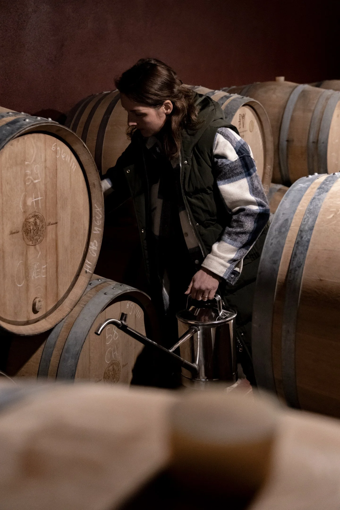 Une femme manipule un tonneau de vin dans une cave à vin, entourée de grands tonneaux en bois.