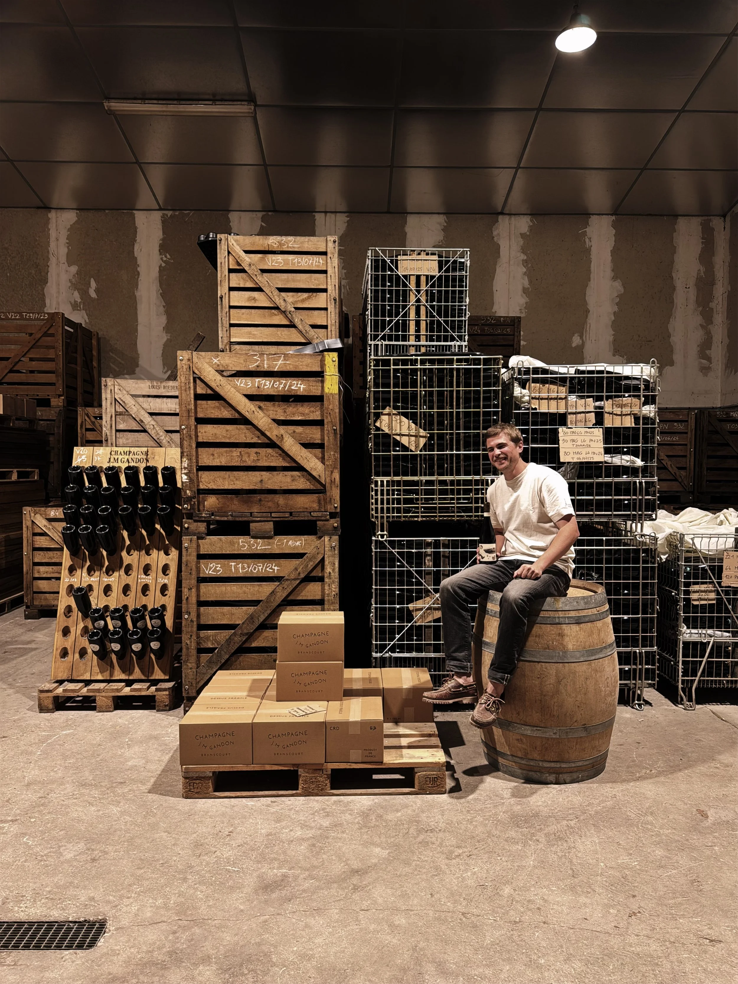 Une personne assise sur un tonneau en bois dans un entrepôt avec des caisses en bois, des cages métalliques contenant des bouteilles, et des cartons empilés, avec un mur en arrière-plan et un plafond en dalles.