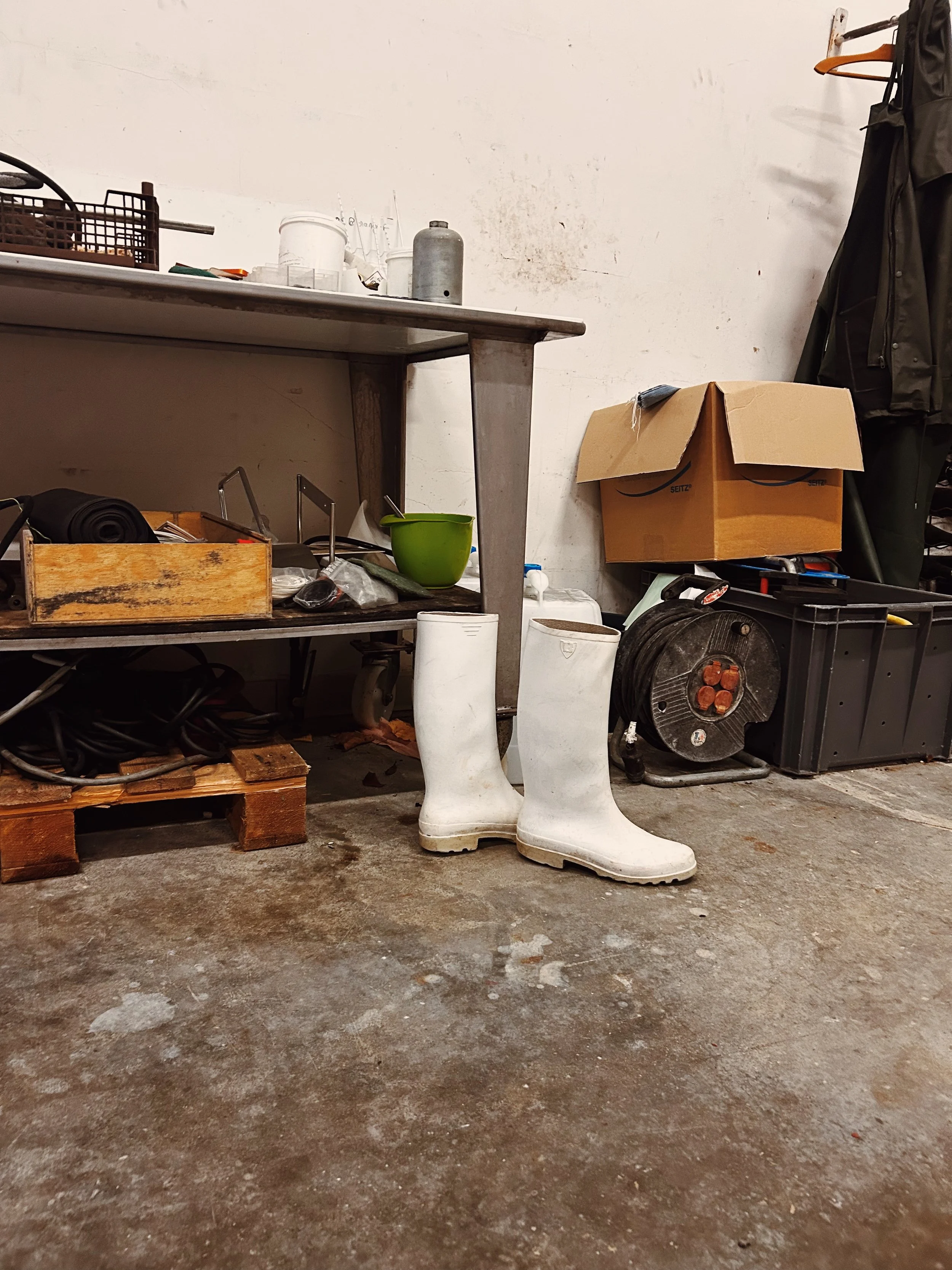 Chaussures en caoutchouc blanc à l'entrée d'un garage ou d'un atelier, avec divers objets et outils posés sur une table et le sol, et un meuble contenant des rouleaux de câble.