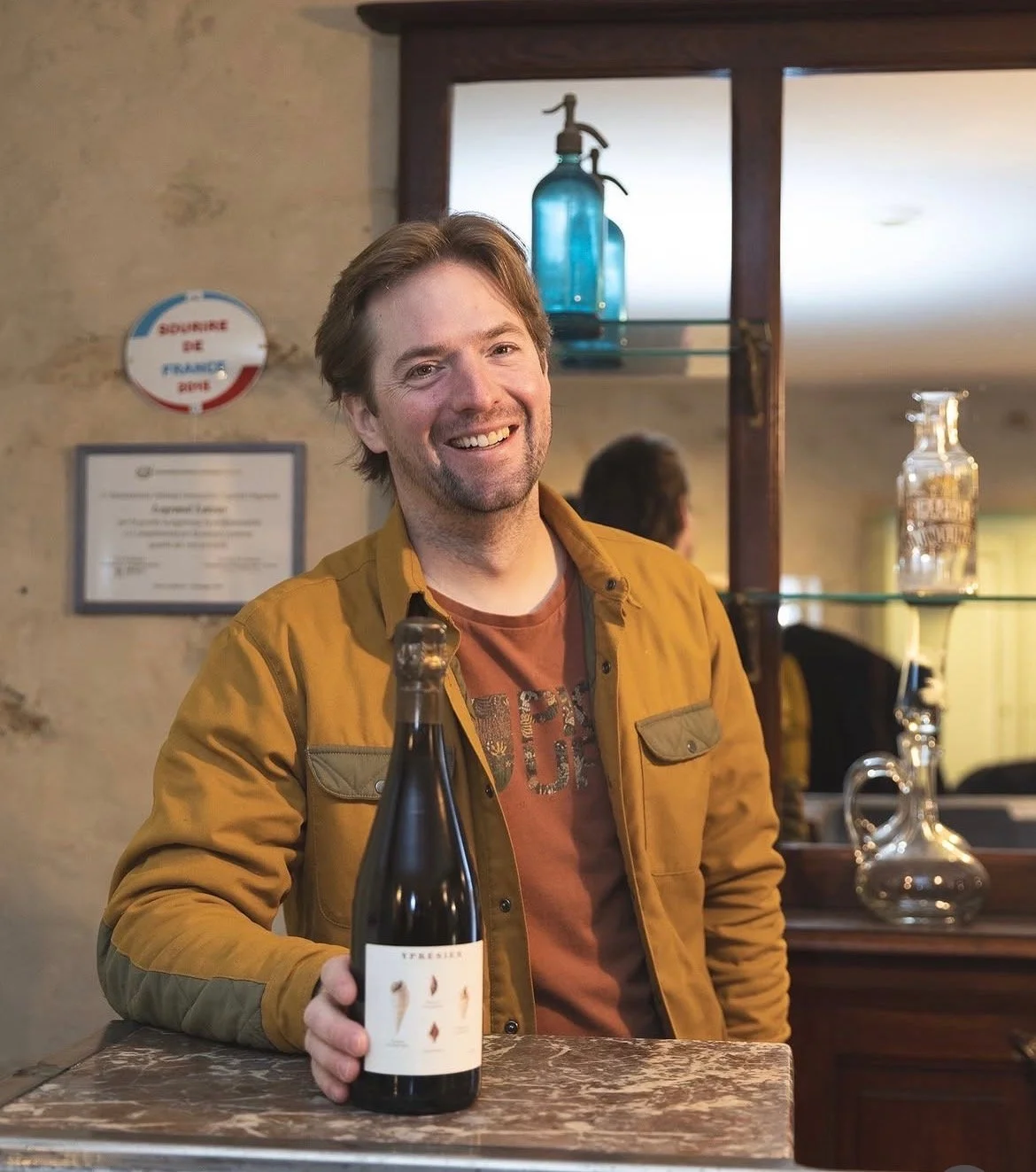 Un homme souriant tenant une bouteille de vin dans un intérieur chaleureux, avec des étagères en verre et des décorations en verre sur fond en pierre.