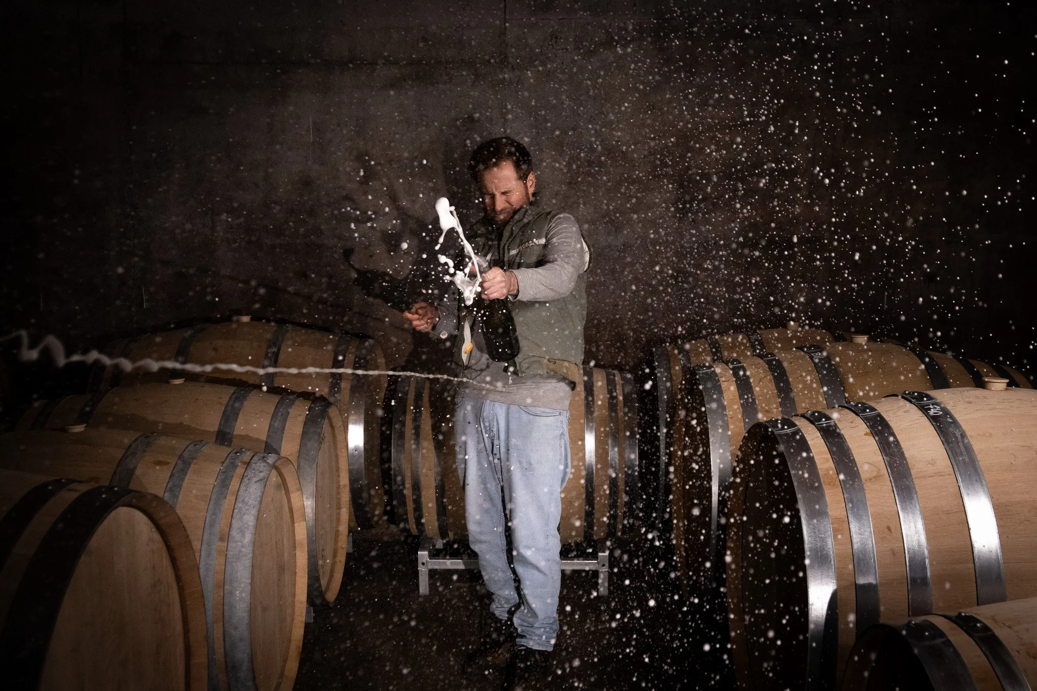 Un homme dans une cave, frappant une barrique de vin avec une baguette, entouré de petites éclaboussures de vin dans l'obscurité.