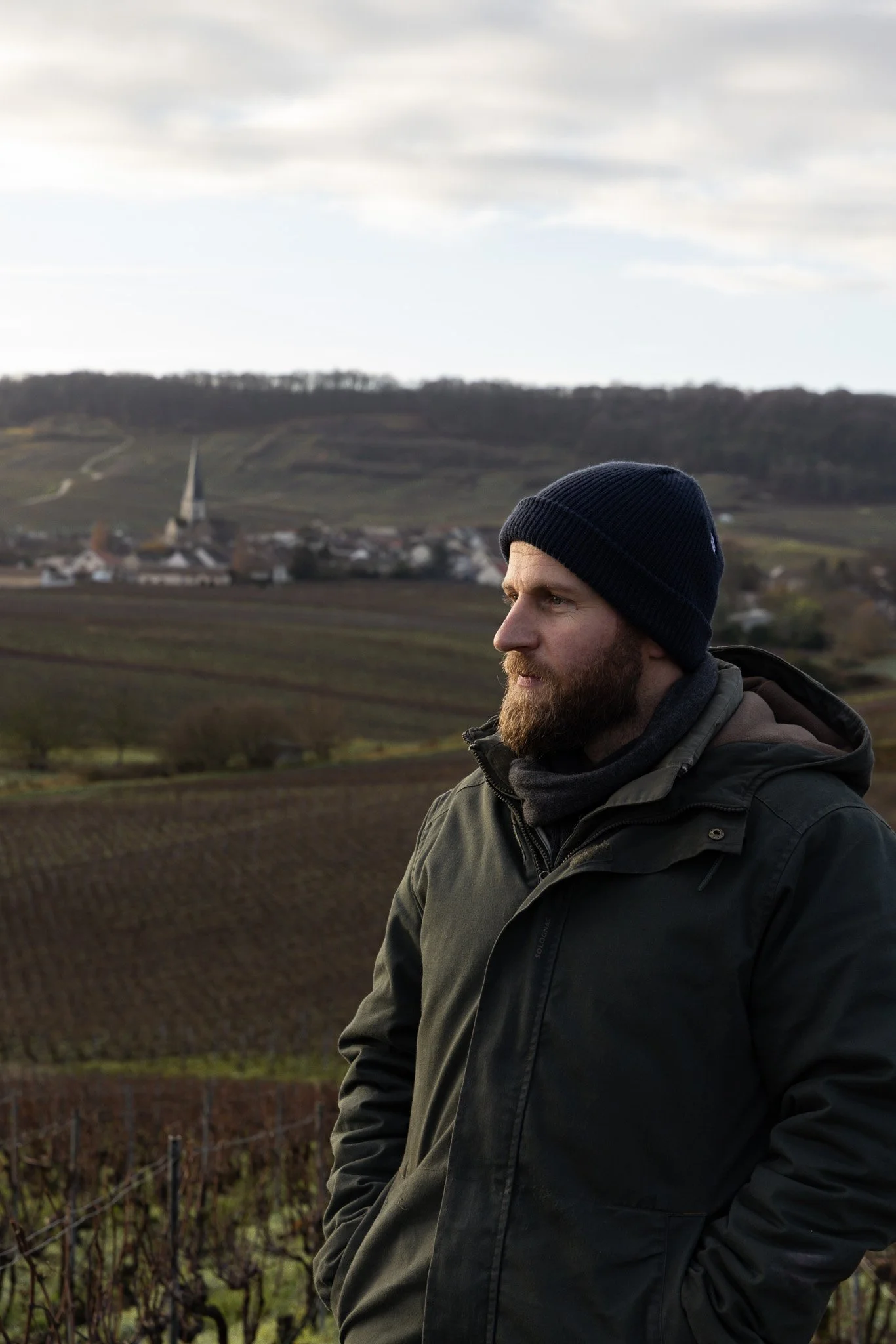 Homme portant un bonnet noir et une veste, regardant vers la droite dans un paysage de campagne avec des vignes et un village en arrière-plan.