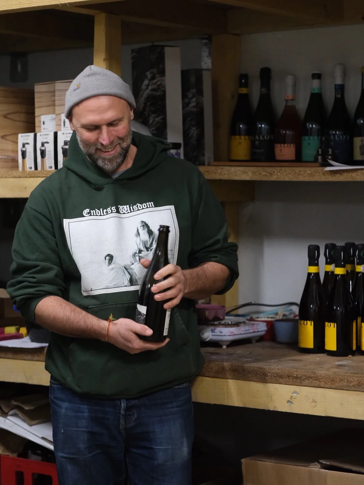 Un homme souriant portant un sweat à capuche vert et une casquette grise, tenant une bouteille en verre noire, dans une pièce avec des étagères de bouteilles et d'autres articles sur le bois
