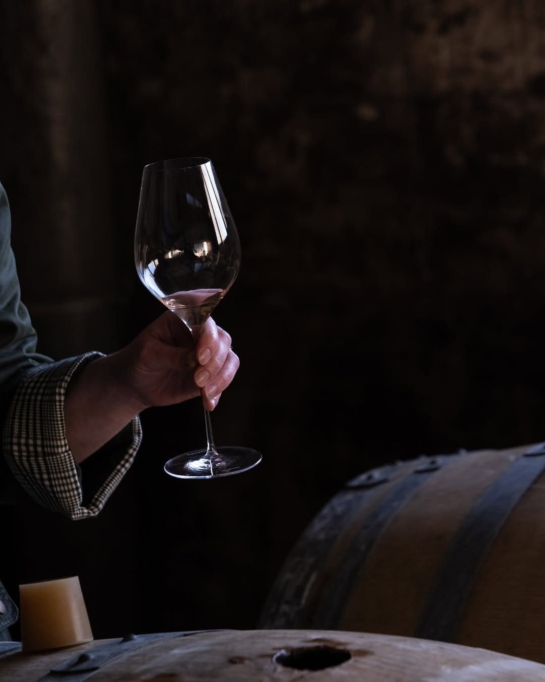 Une personne tient un verre à vin vide en main, dans une cave sombre avec des tonneaux en bois.