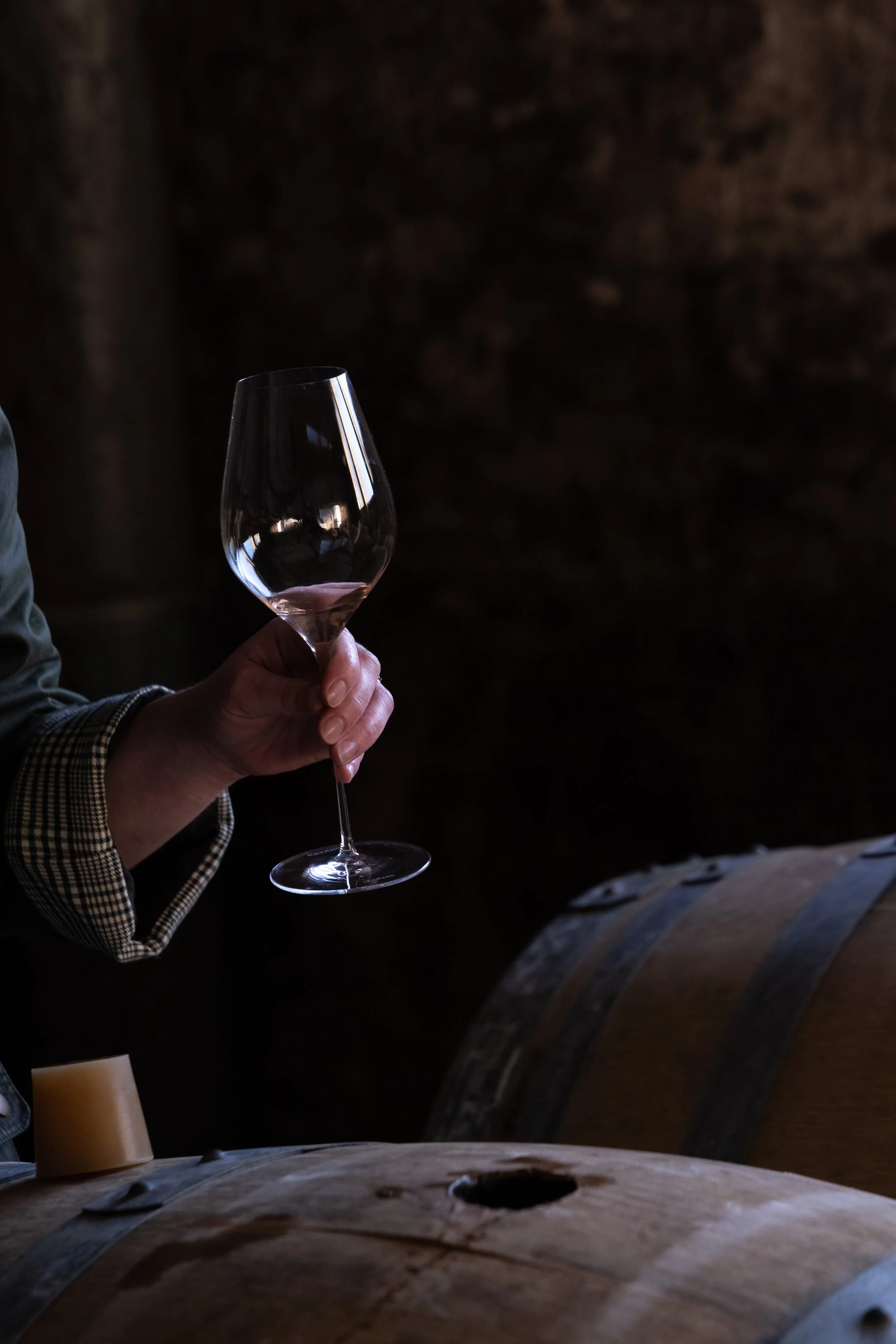 Une personne tient un verre à vin vide dans un environnement sombre, avec un grand tonneau en bois en arrière-plan.