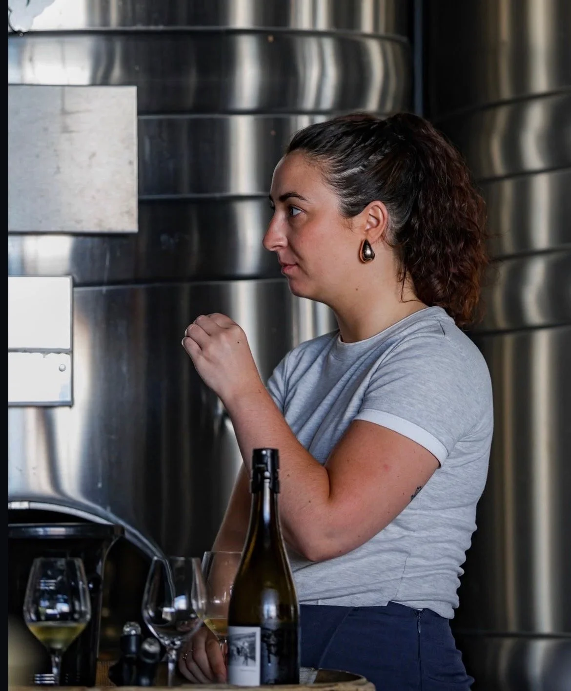 Une femme avec des cheveux bouclés attachés en arrière, portant un t-shirt gris, prépare une dégustation de vin dans une cuisine en acier inoxydable.