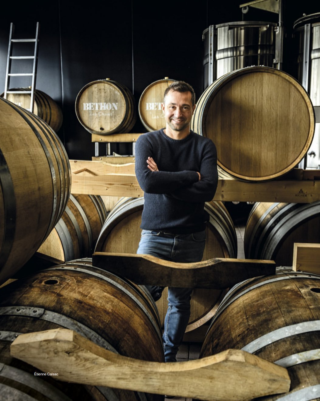 Un homme souriant avec des bras croisés dans une cave à vin, entouré de fûts en bois, dans un environnement de cave à vin