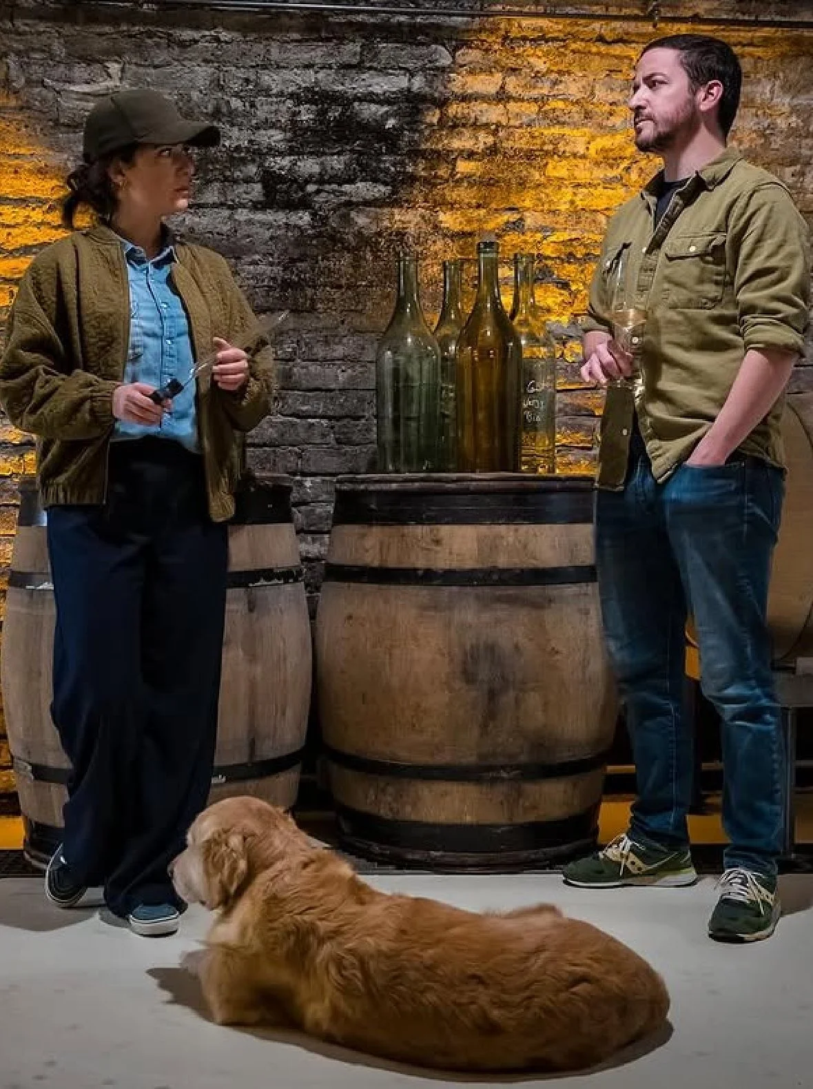 Deux personnes discutent dans une cave à vin avec des bouteilles de vin en verre sur une table, un chien lazar repose au sol, et des tonneaux en bois sont visibles.