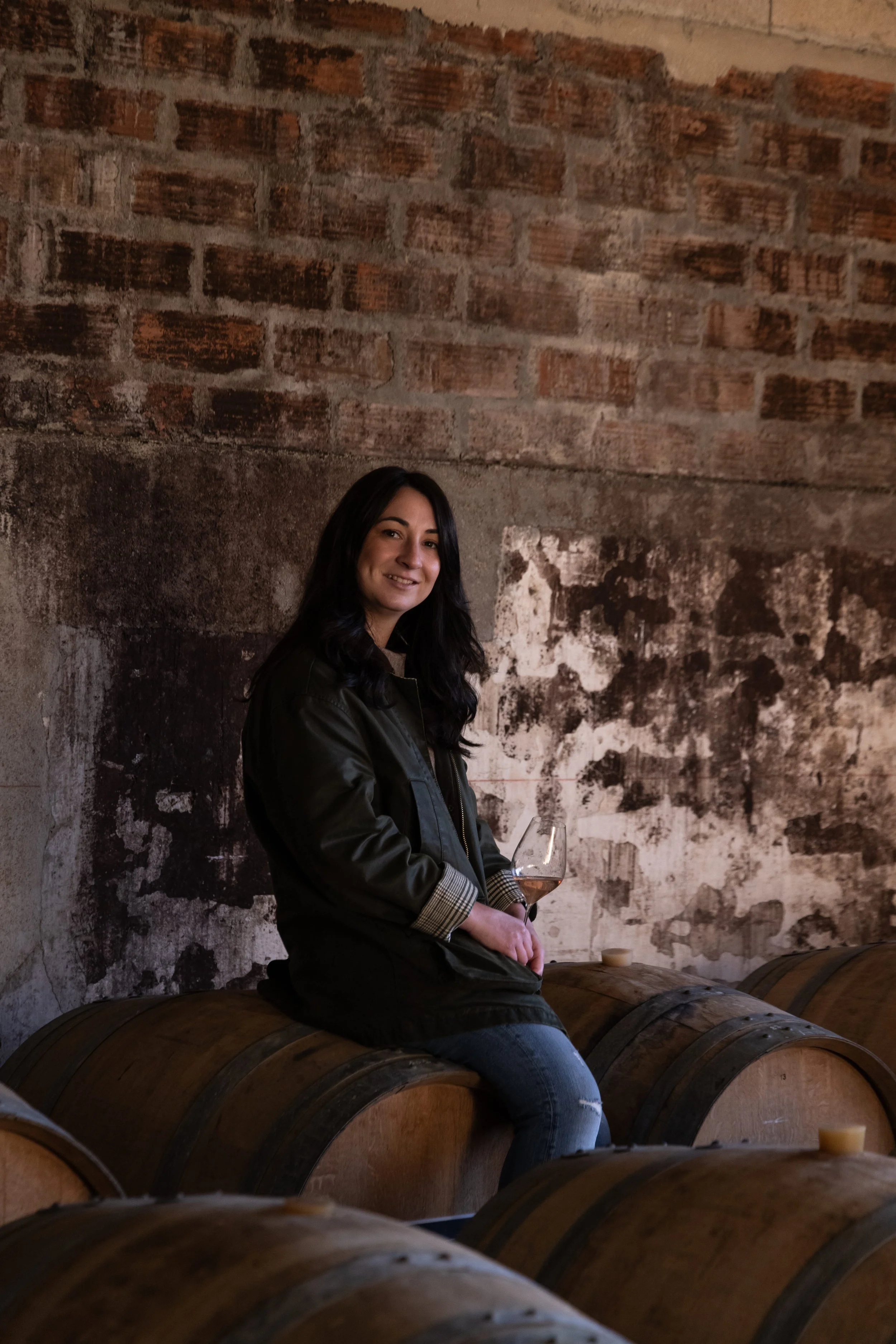 Femme assise sur des tonneaux en bois dans un endroit avec un mur en briques et en plâtre, tenant un verre de vin rosé.