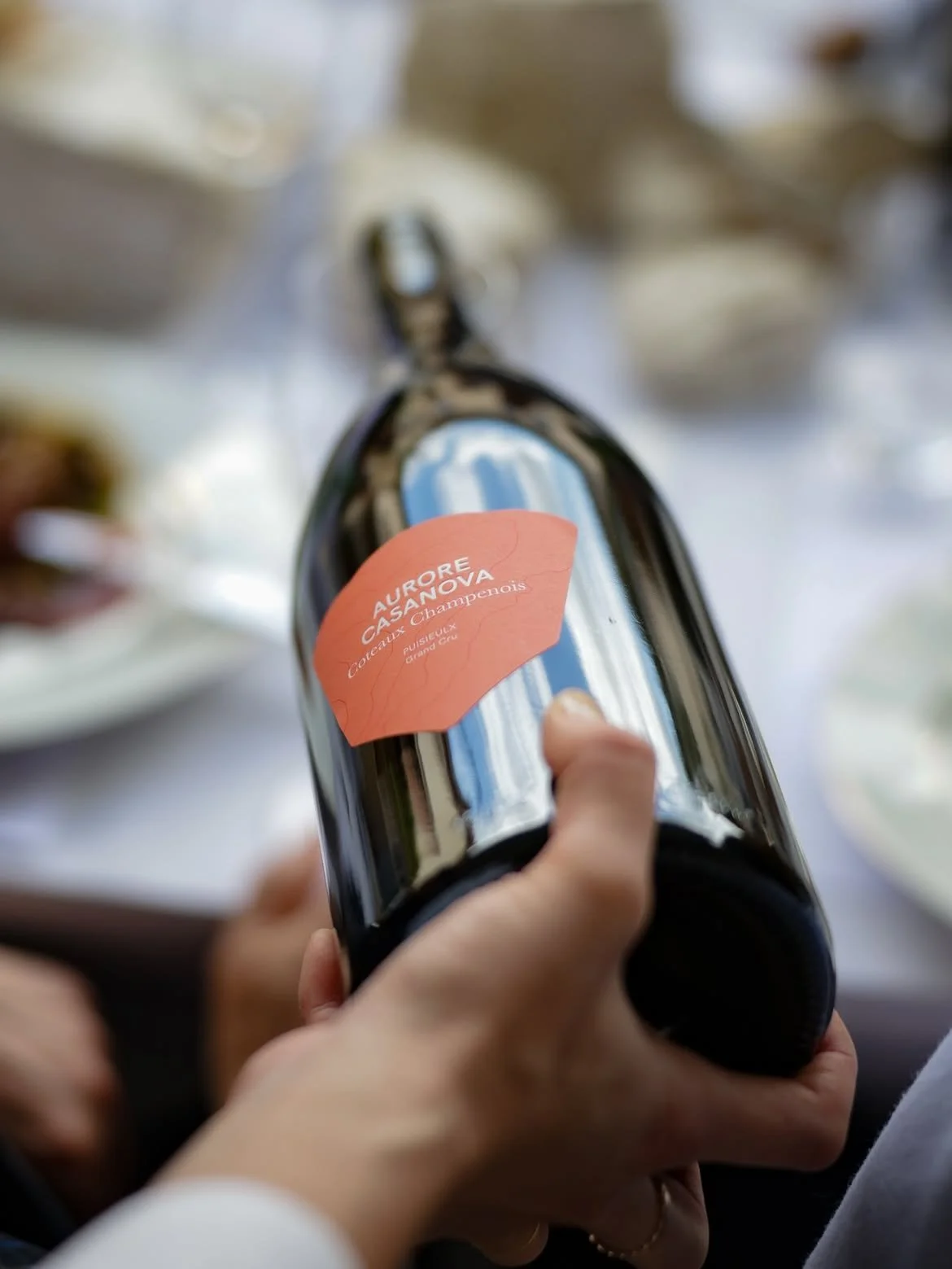 Une personne tient une bouteille de vin avec une étiquette orange indiquant 'Aurora Casanova, Coteaux Champenois, pommesulx et cru' contre un fond flou.