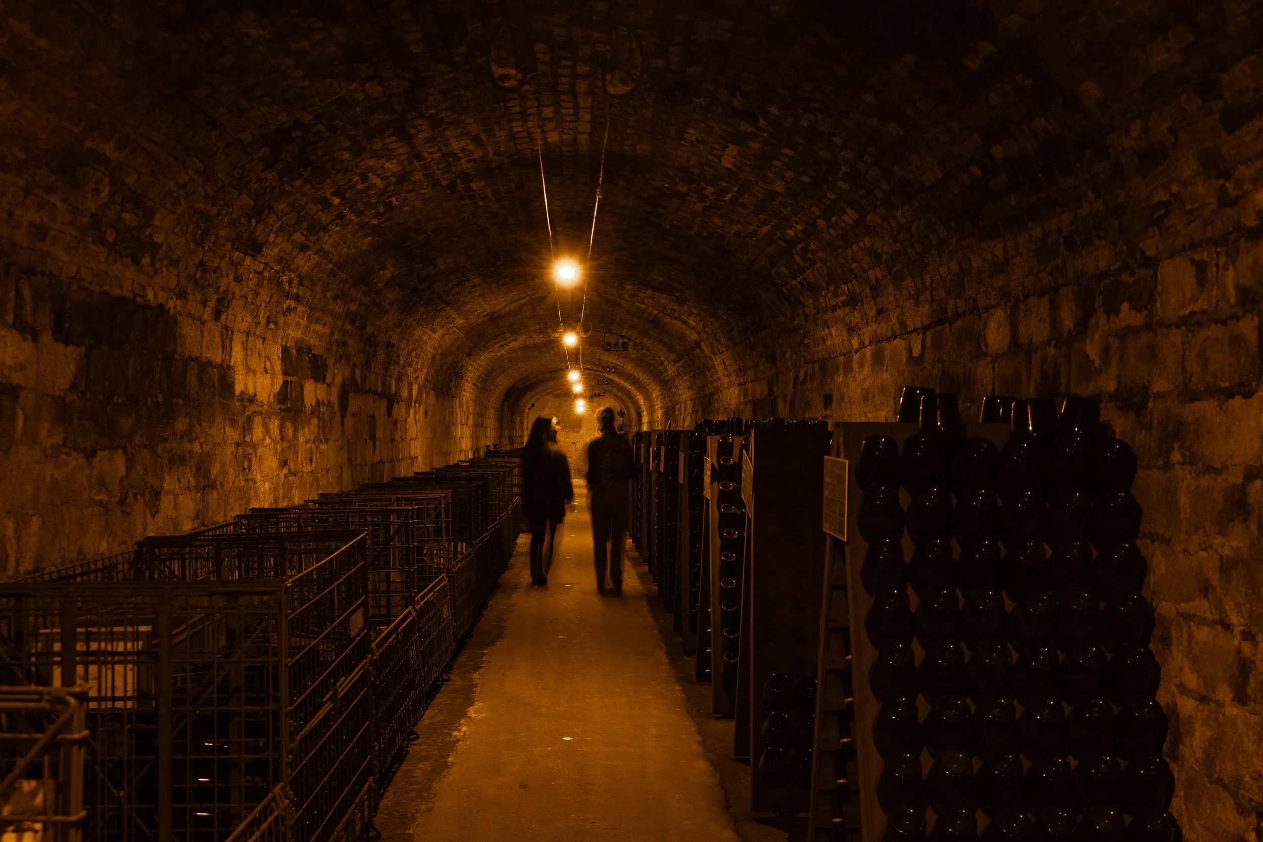 Intérieur d'une cave ou d'un tunnel en pierre, éclairé par des lampes suspendues. Deux personnes discutent au centre, entourées de charriots et de bouteilles de vin.
