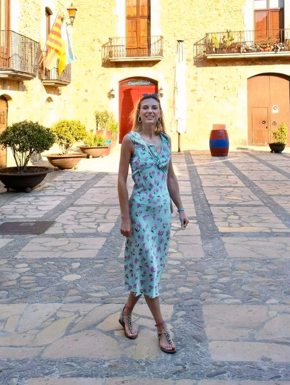 A woman with shoulder-length hair, wearing a sleeveless light blue floral dress and sandals, walking on a cobblestone plaza with potted plants and a historic yellow brick building in the background.