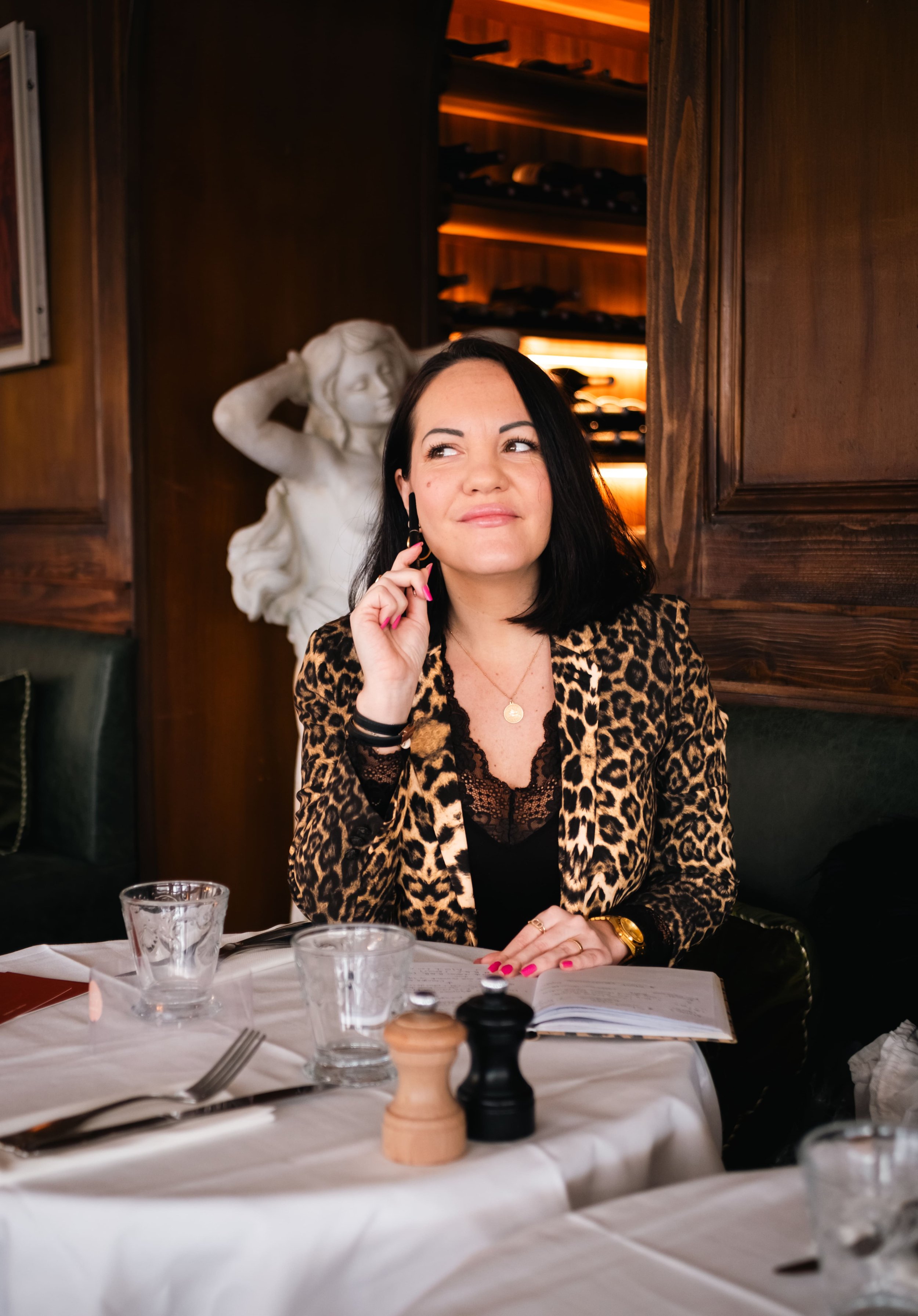 Femme avec cheveux noirs, portant un blazer à motif léopard, assise à une table dans un restaurant, en train de réfléchir ou de sourire légèrement, avec une sculpture de femme en arrière-plan.