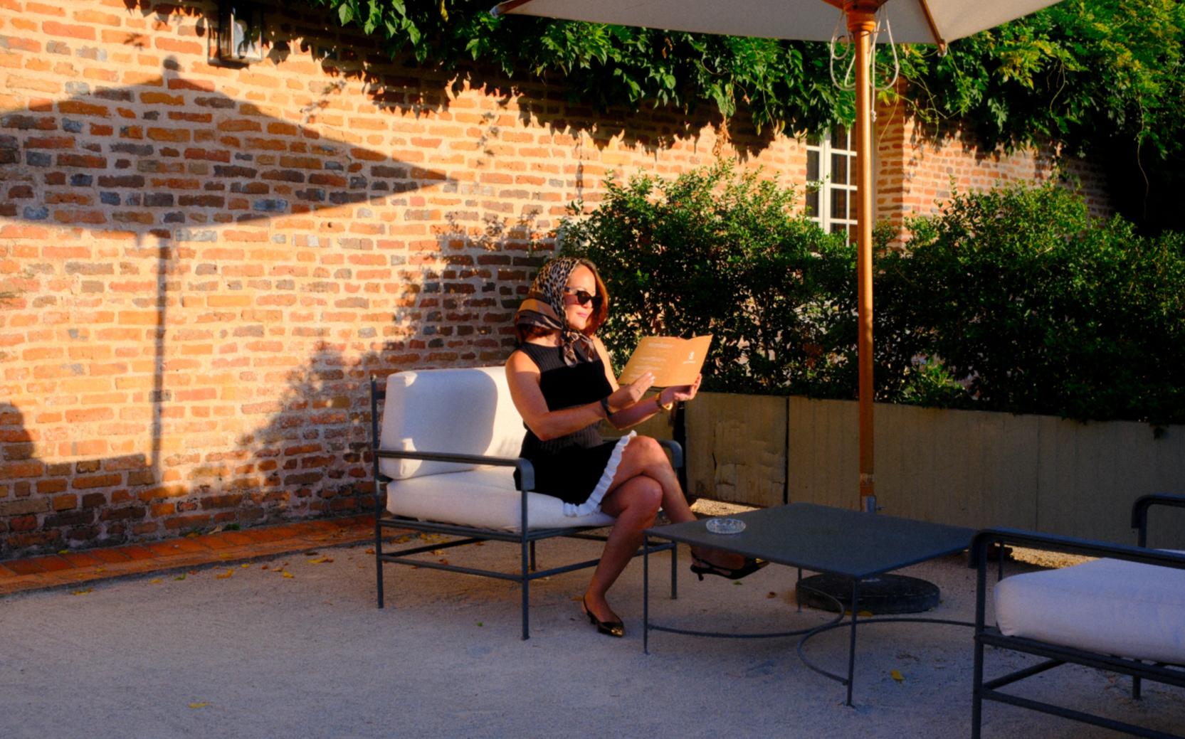 Une femme assise sur un fauteuil extérieur, lisant un menu ou un livre, sous un parasol avec une véranda en briques derrière elle, entourée de buissons.