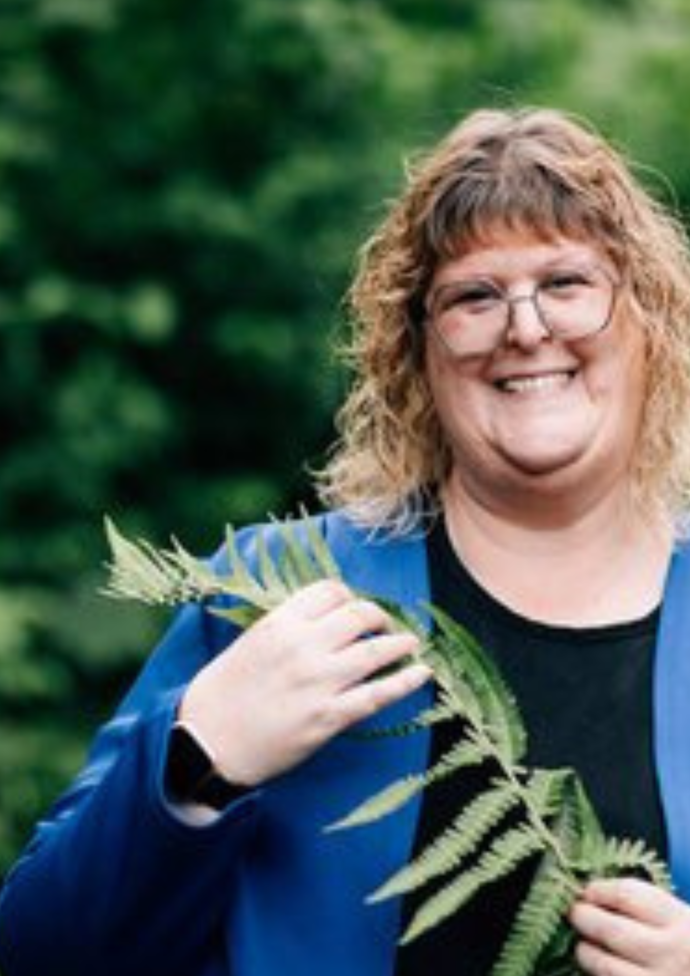 Eine lächelnde Frau mit lockigem, braunem Haar und Brille hält einen Farnwedel in der Hand. Diese Frau bin ich.
