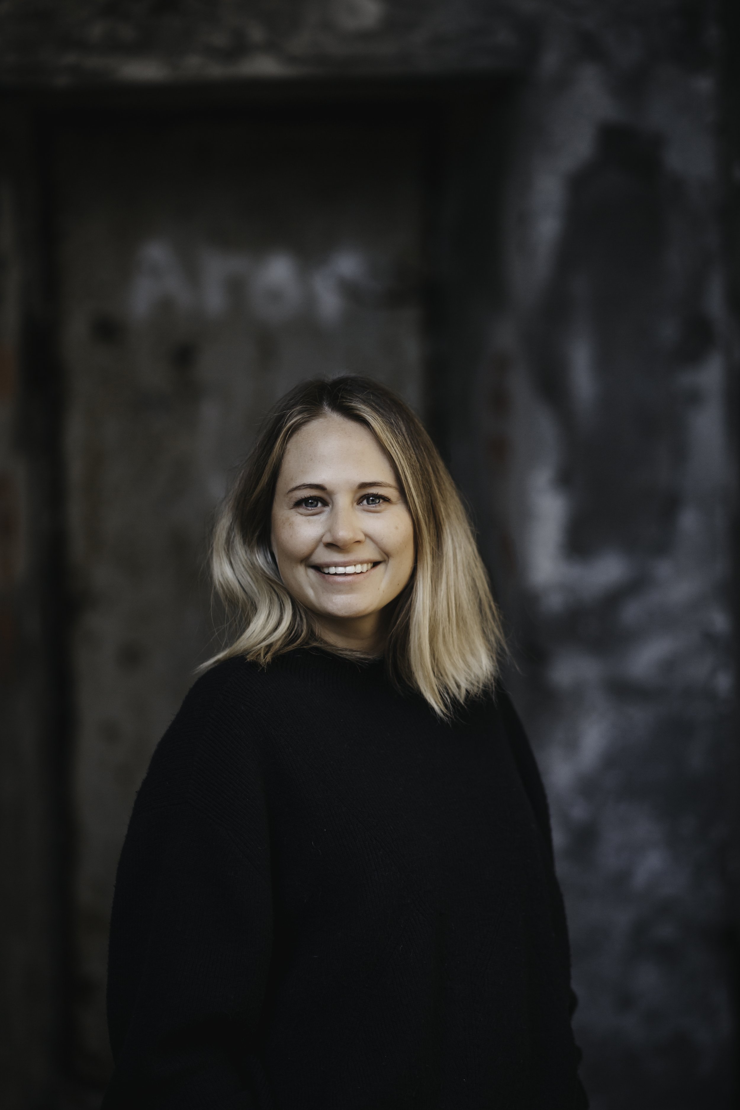 Close-up of a smiling woman with blonde hair wearing a black sweater, standing against a dark, textured background with graffiti.