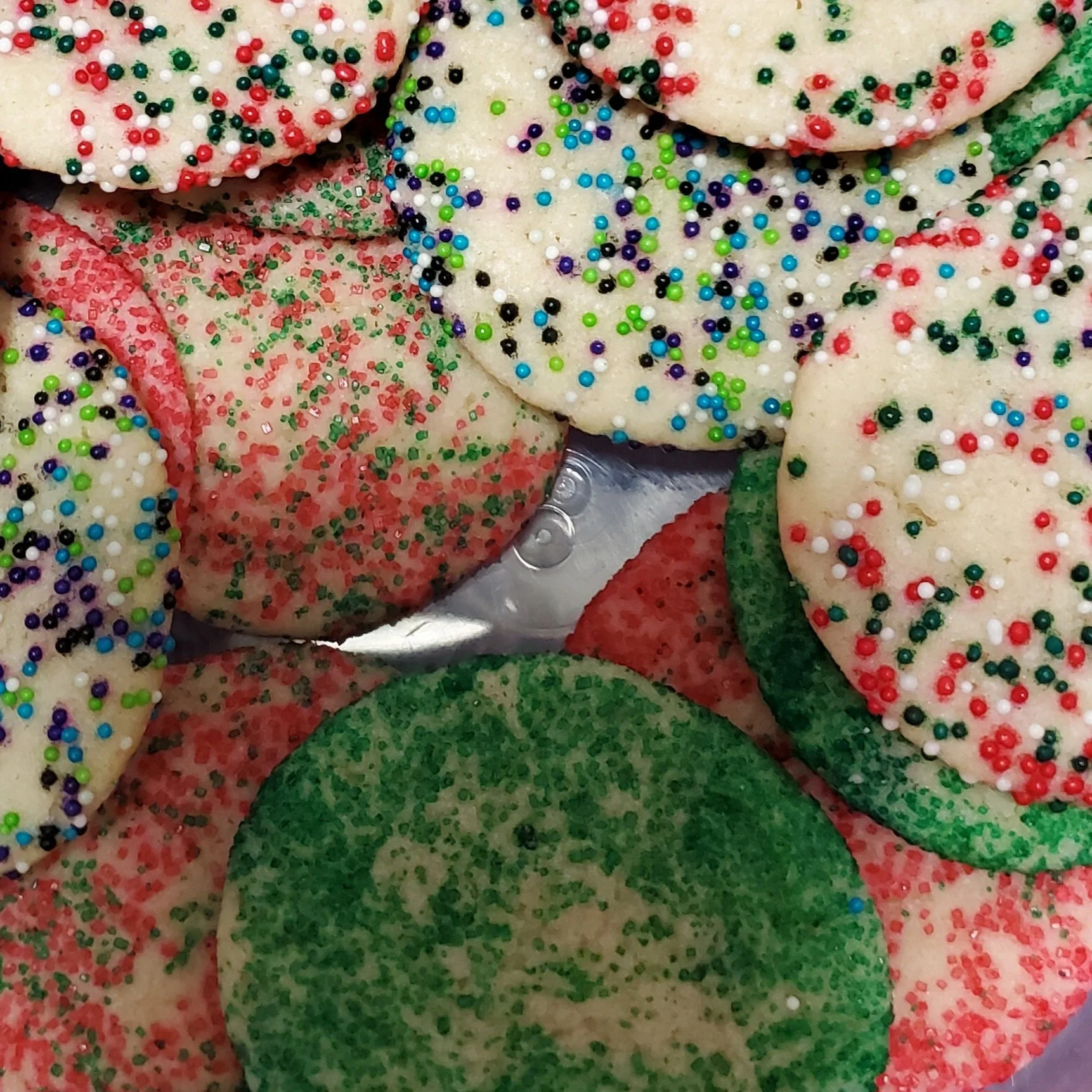 Close-up of sugar cookies decorated with assorted colorful sprinkles, including red, green, blue, purple, white, and black, shaped as snowflakes, stars, and round beads.