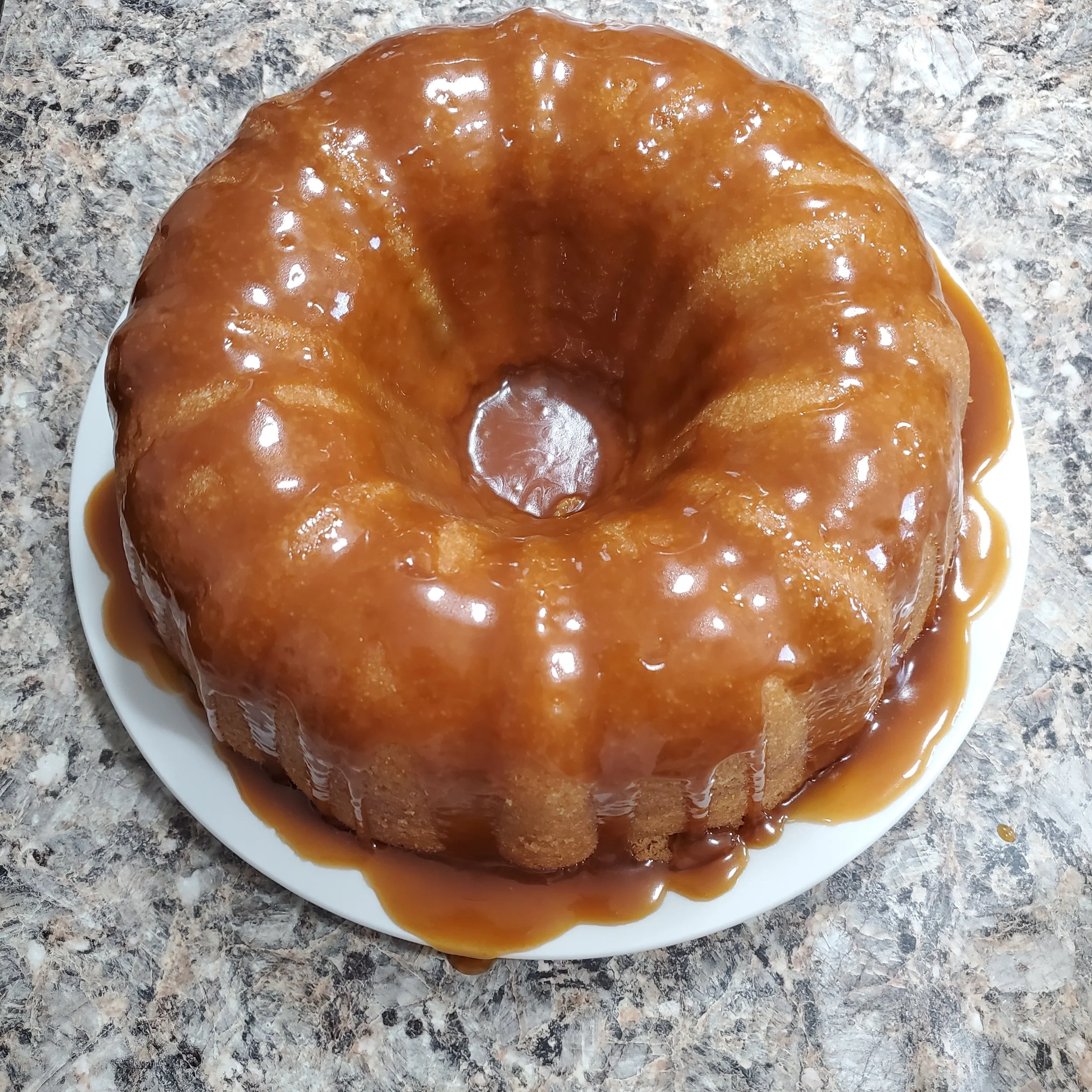 A caramel Bundt cake with caramel glaze on a white plate, placed on a speckled granite countertop.