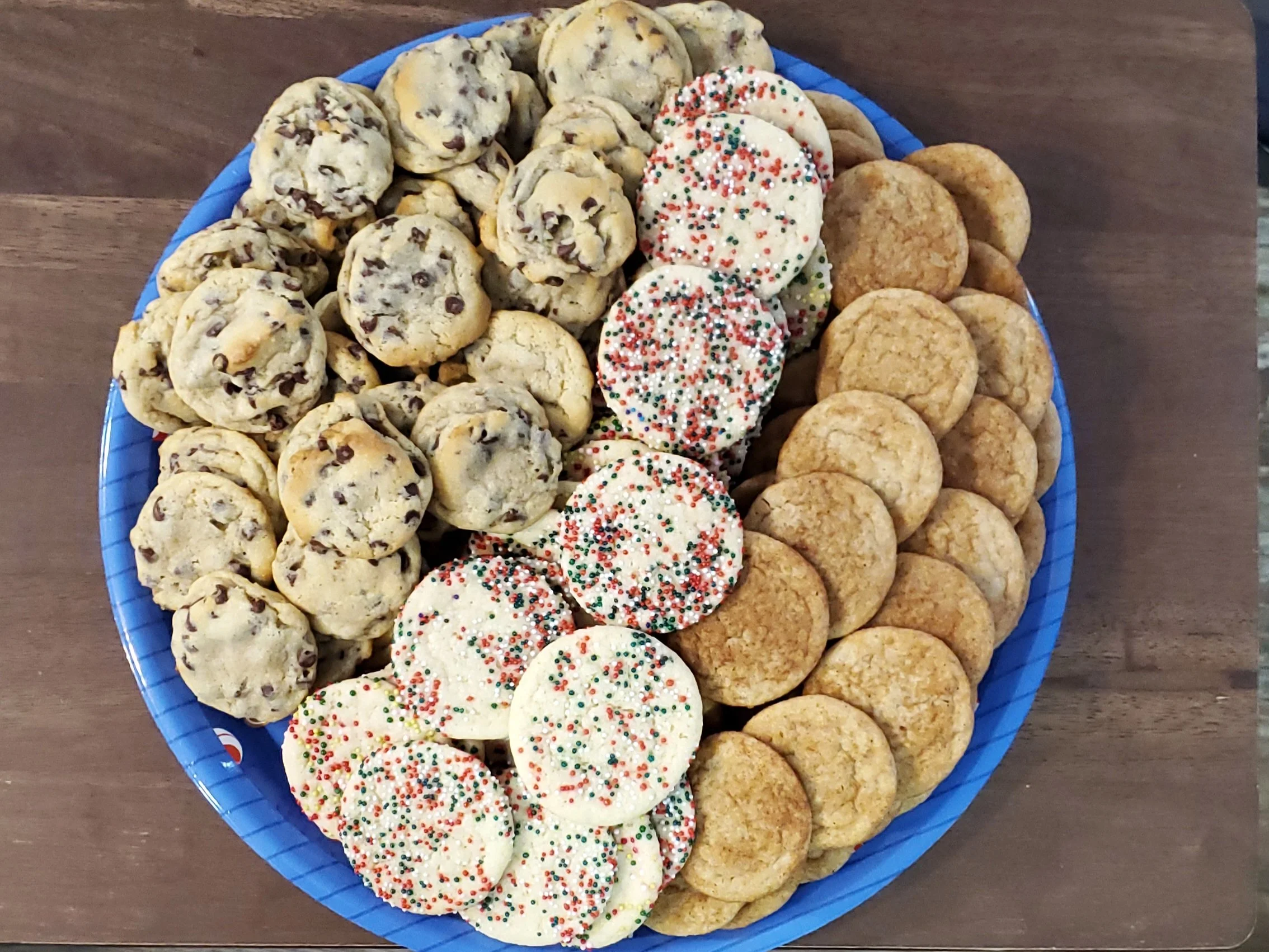 A blue plastic plate with three types of cookies: chocolate chip cookies, sugar cookies with colorful sprinkles, and peanut butter cookies, placed on a wooden surface.