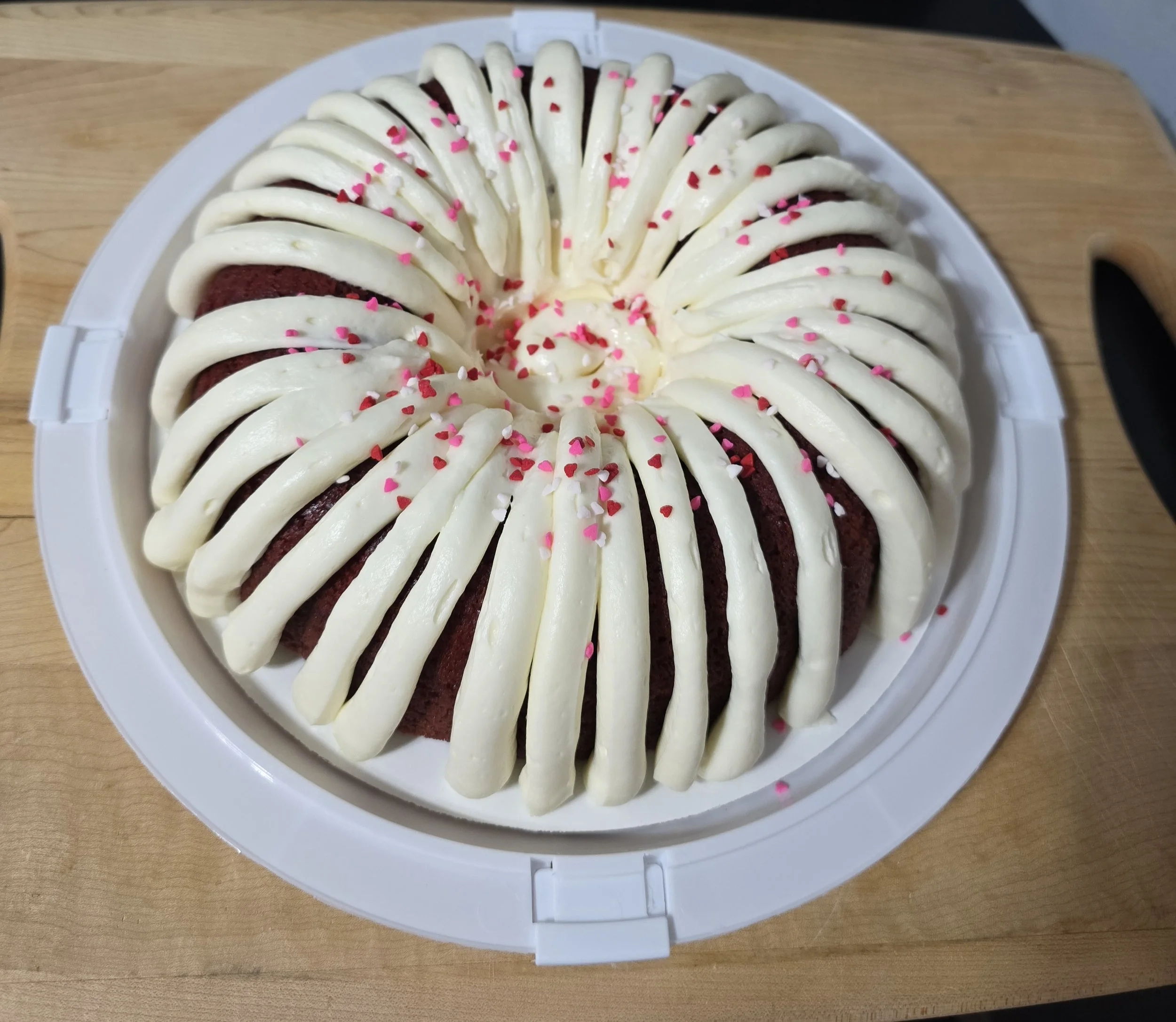 A round cake decorated with white frosting, pink and red heart-shaped sprinkles, and white chocolate curls arranged in a circular pattern on top.