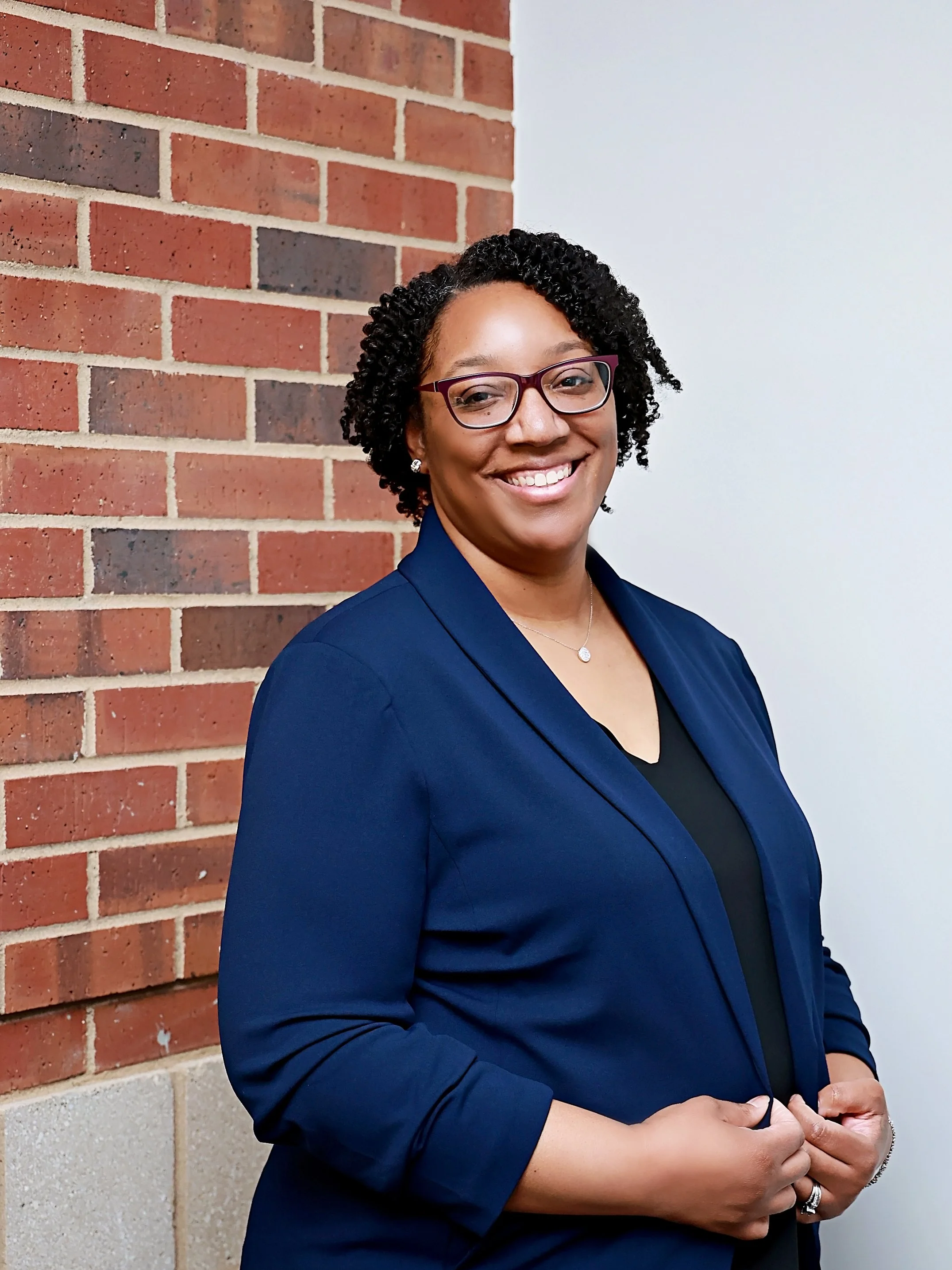 A woman with curly black hair wearing glasses, a dark blazer, and a necklace, smiling, standing near a brick wall and a white wall.