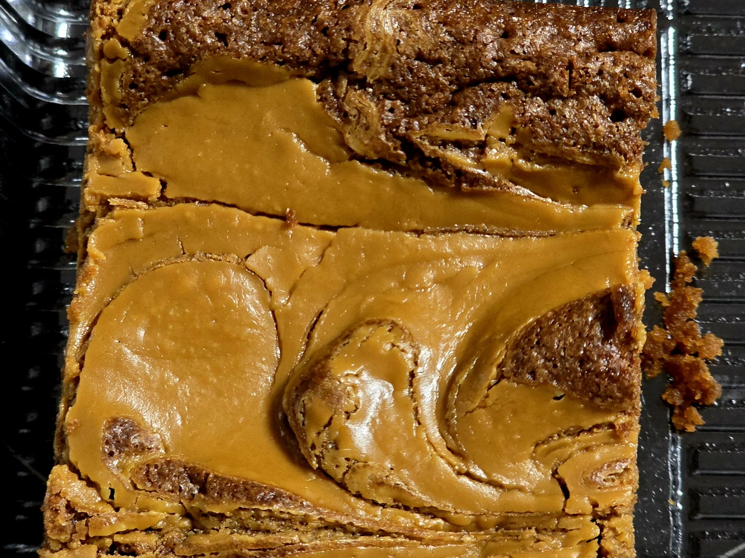 Close-up of a square piece of layered dessert with a yellow caramel topping and a brown crumbly layer beneath, on a black wire rack.
