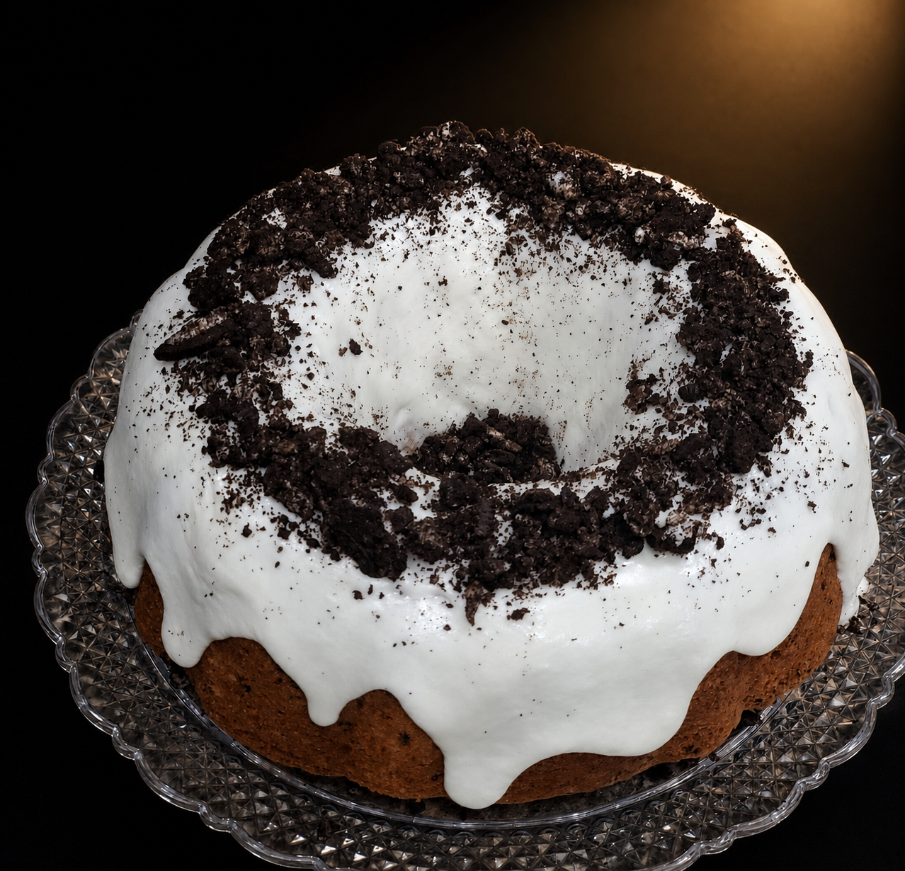 Chocolate bundt cake with white icing and crushed cookies on top served on a glass platter.