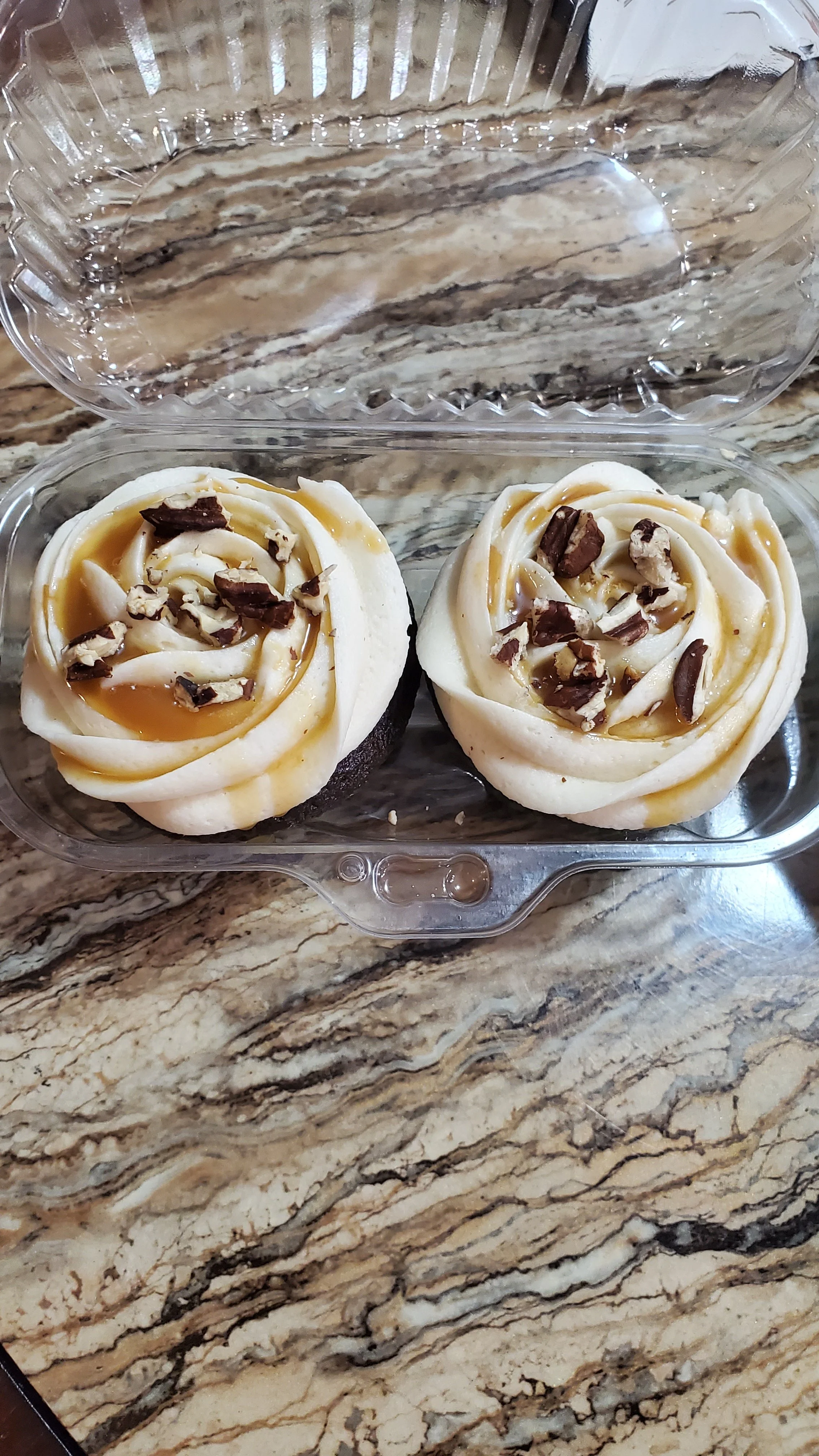 Two cupcakes decorated to look like roses with white frosting, caramel drizzle, and chopped chocolate on top, in a clear plastic container on a marble countertop.