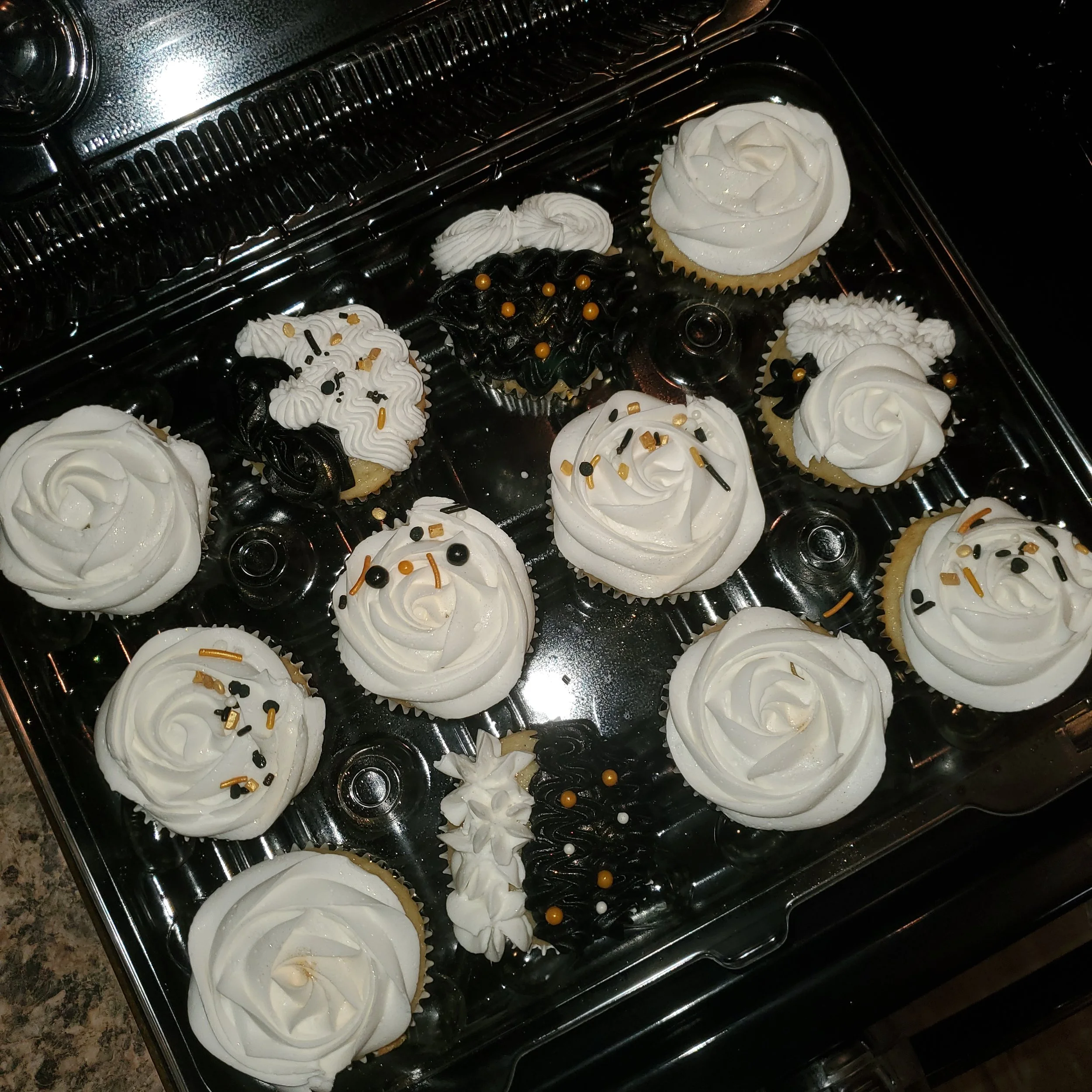Assorted cupcakes in a clear plastic container, decorated with white, black, and gold sprinkles and icing with swirls and rosettes.