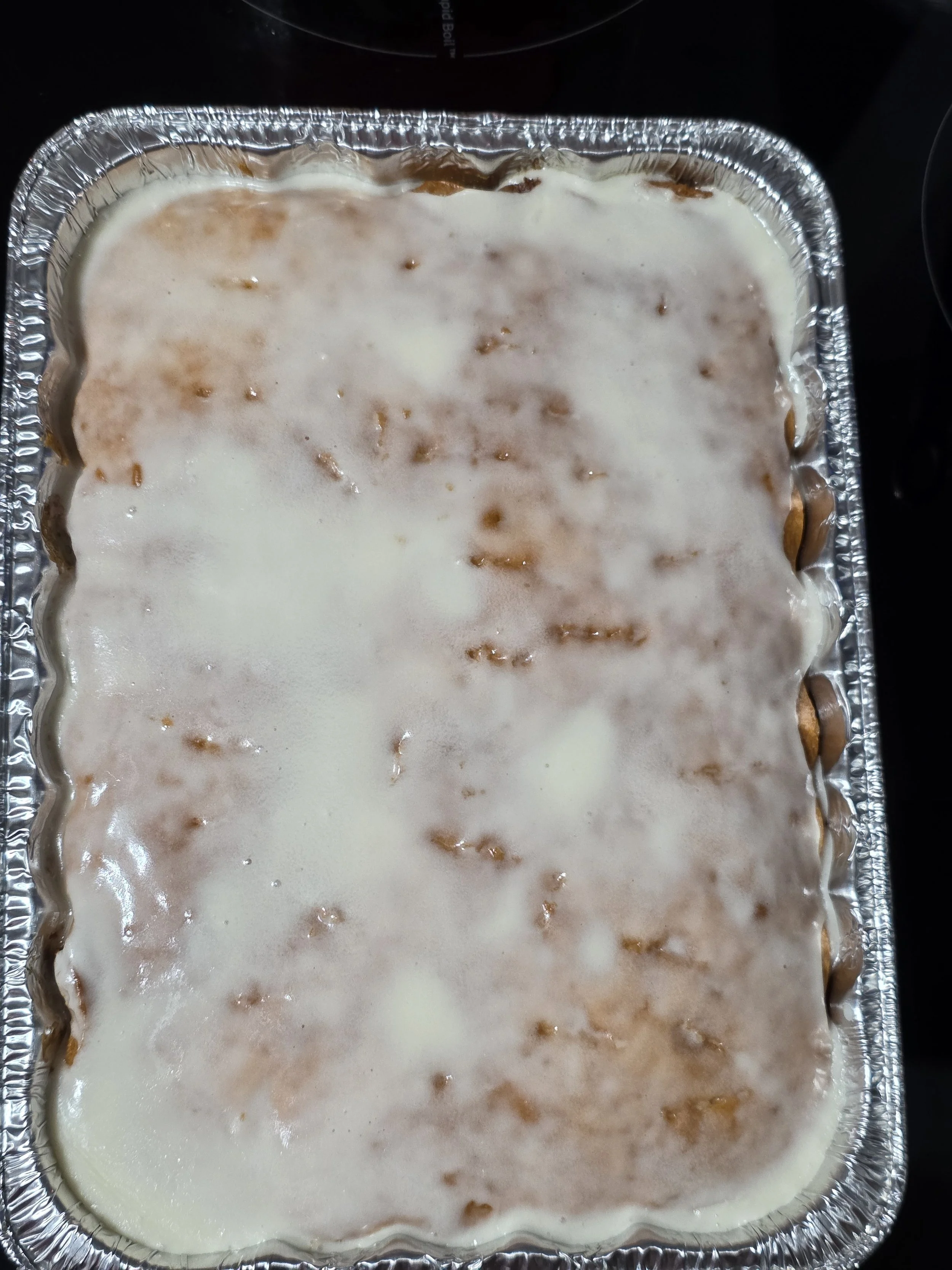 Aluminum container with a layered dish topped with a white sauce or frosting and some brown crumbs or pieces underneath. The dish appears to be a baked casserole or dessert.