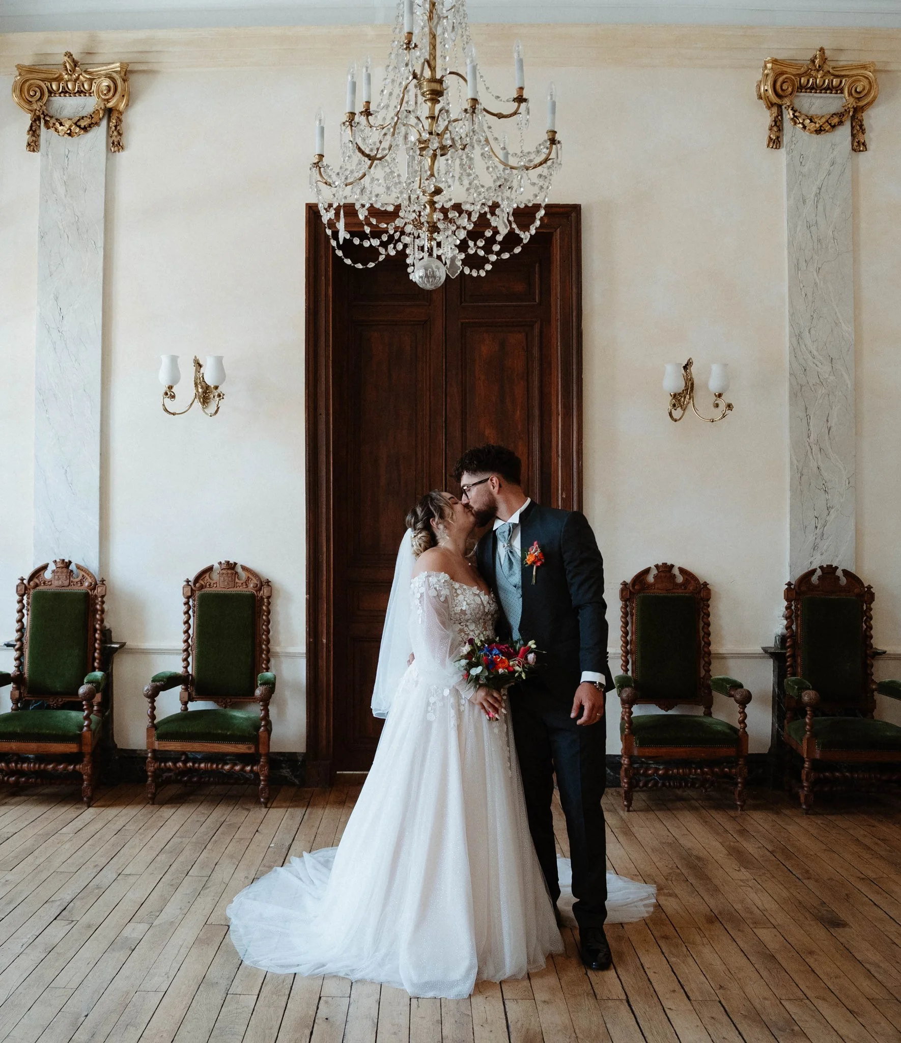 Un couple marié s'embrasse dans une salle élégante avec un chandelier en cristal suspendu et des fauteuils en bois avec des coussins verts.