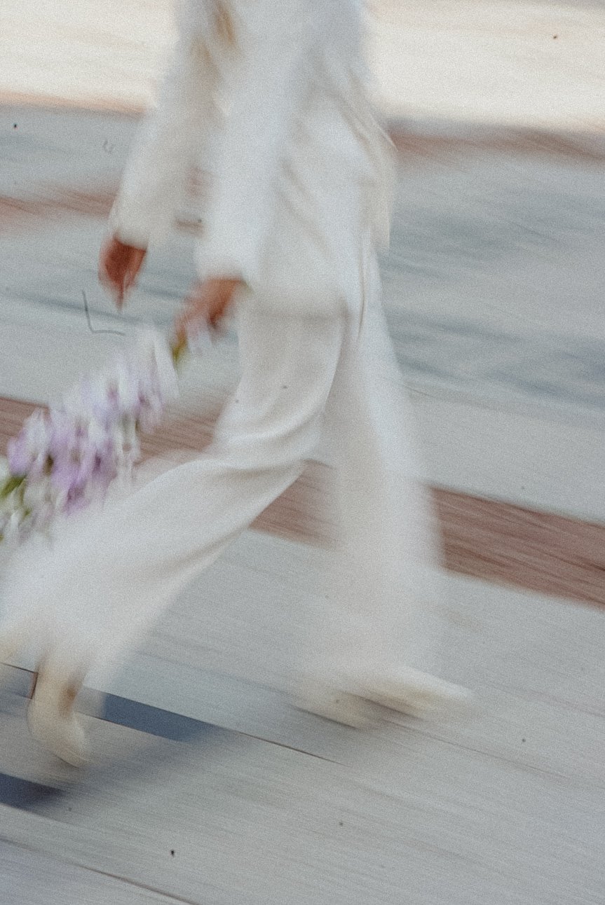 Une personne en robe blanche avec des chaussures blanches, tenant un bouquet de fleurs violettes, marche sur un sol en bois, dans une image floue et en mouvement.